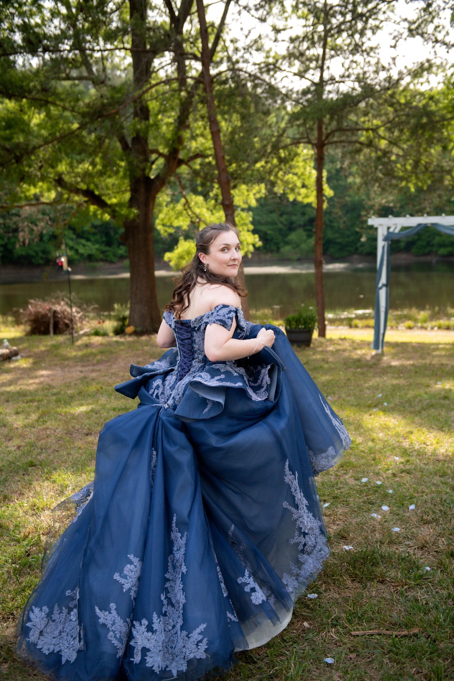Young woman twirling in an elegant navy blue off-shoulder ball gown with lace trim outdoors near a pond.