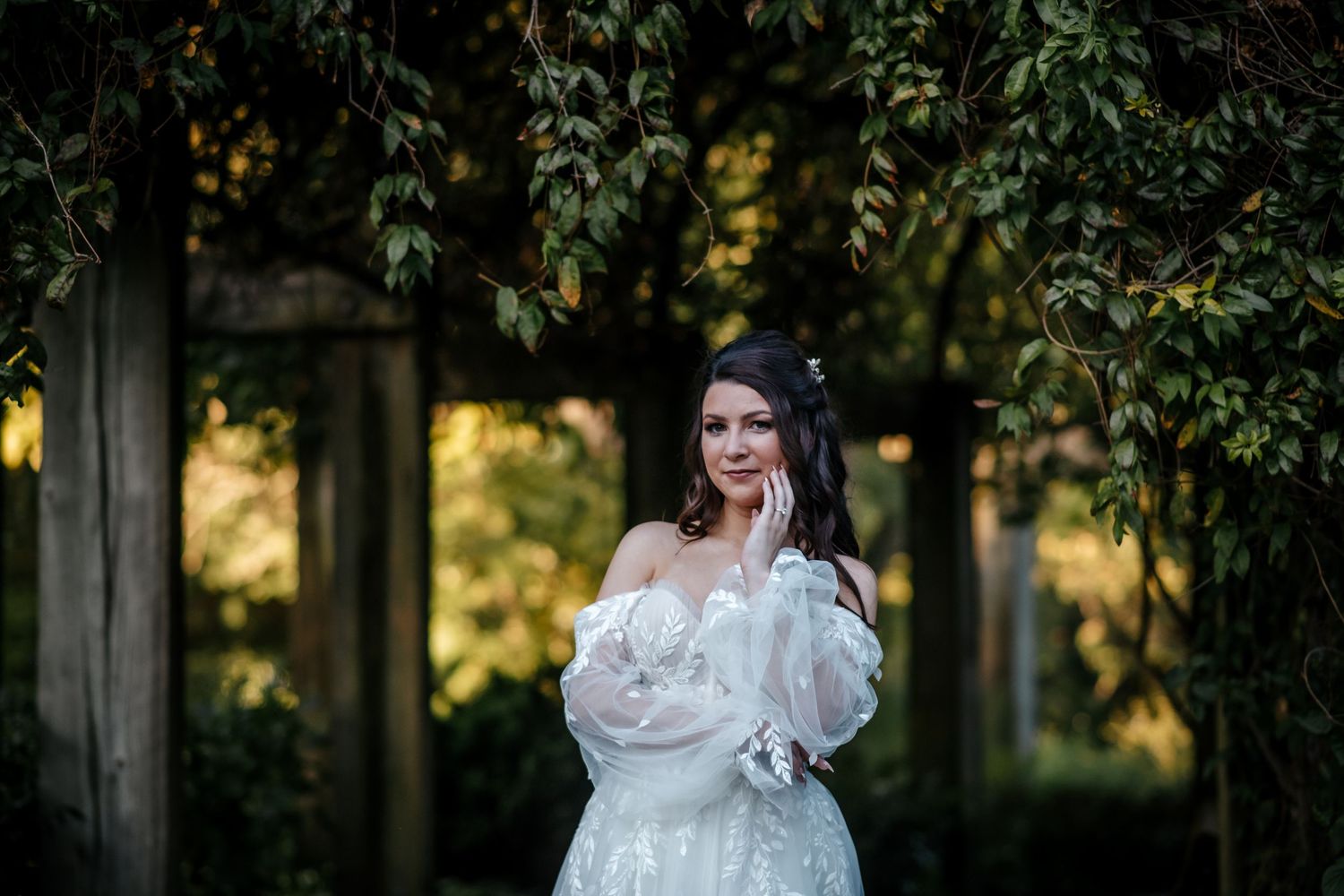 Bride in white off-shoulder gown poses gracefully among lush green ivy-covered wooden pillars in a garden.