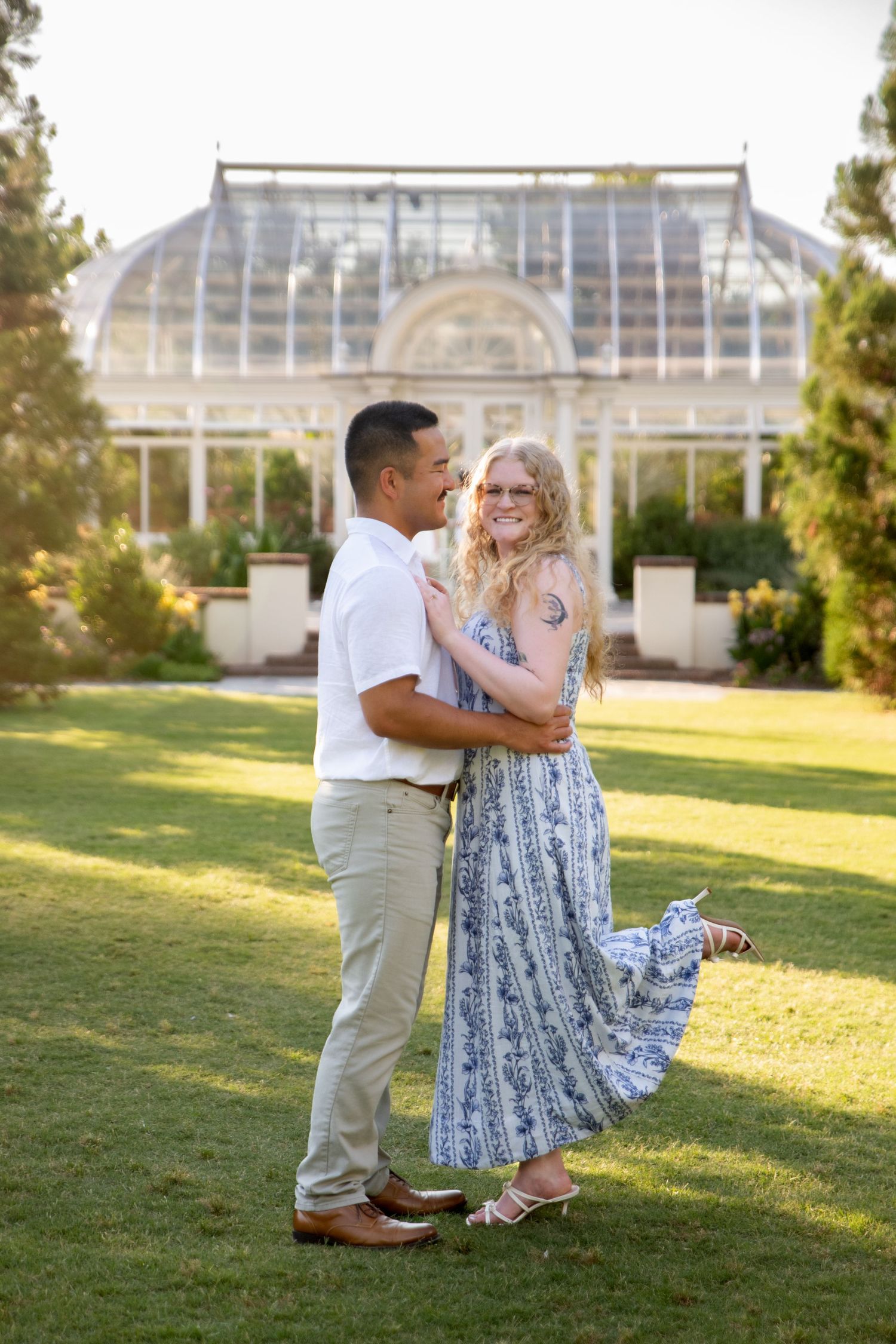 Couple embracing and smiling in front of a glass greenhouse in a lush green garden, woman in blue floral dress.