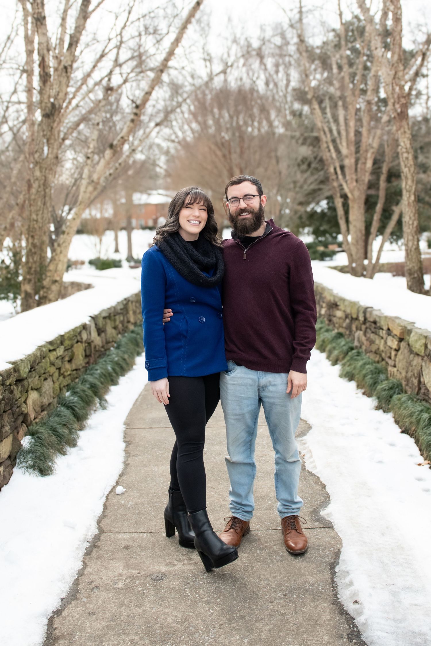 A couple in winter attire poses playfully on a snowy path lined with evergreen trees.