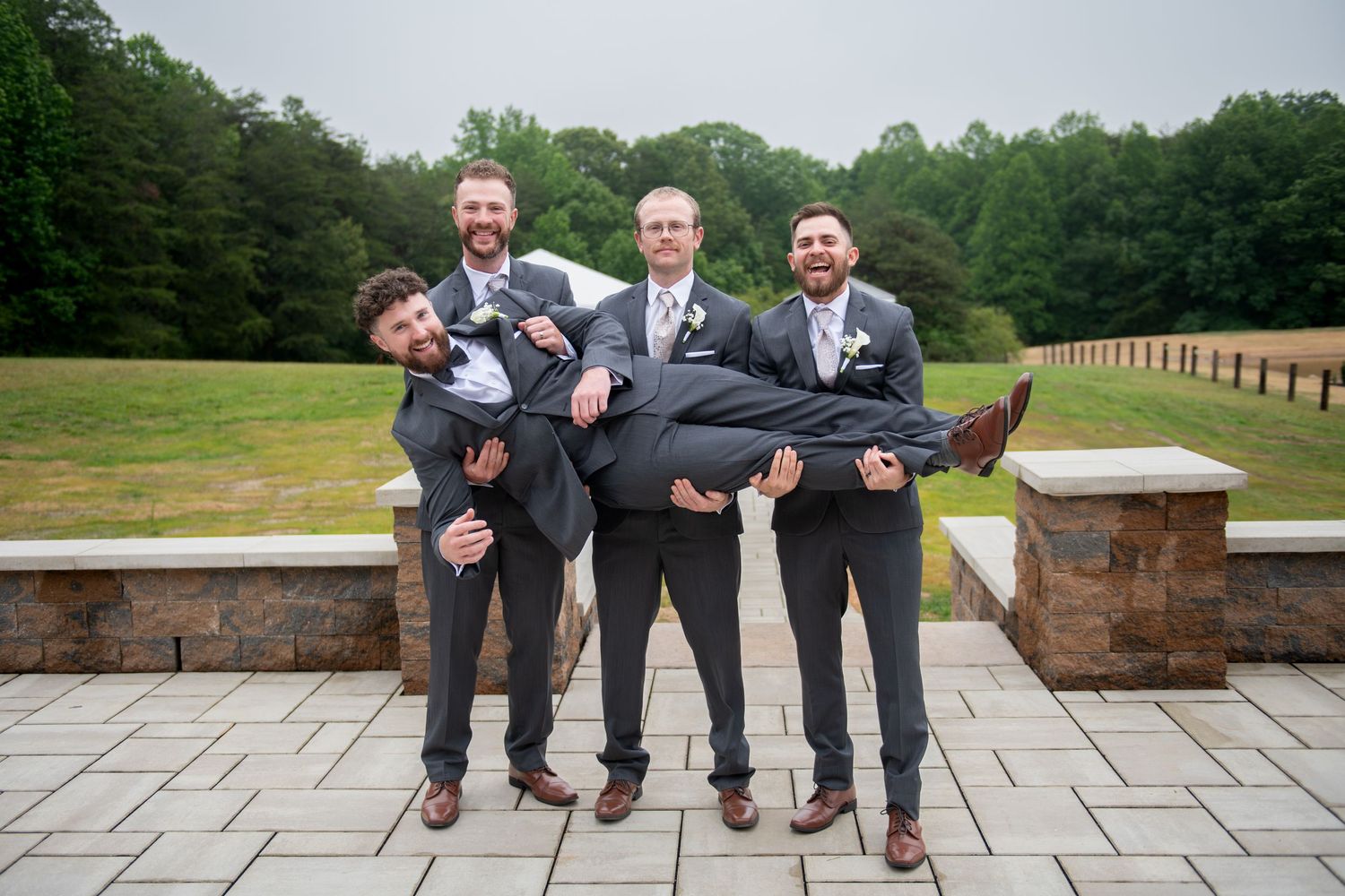 Groomsmen in gray suits lifting the groom outdoors on a stone patio with lush green trees in the background.