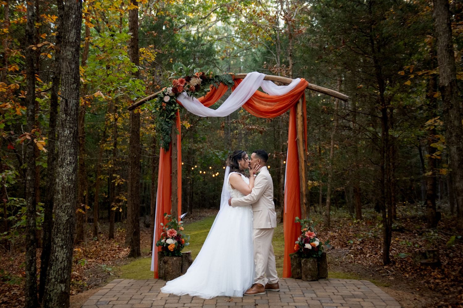 Bride and groom kissing under a rustic floral arch with orange draping in an autumn forest wedding ceremony.