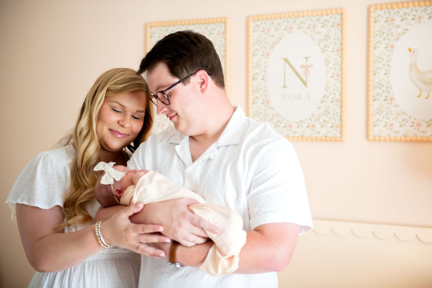 New parents lovingly cradle their newborn baby in a softly decorated nursery with framed letter art on the wall.
