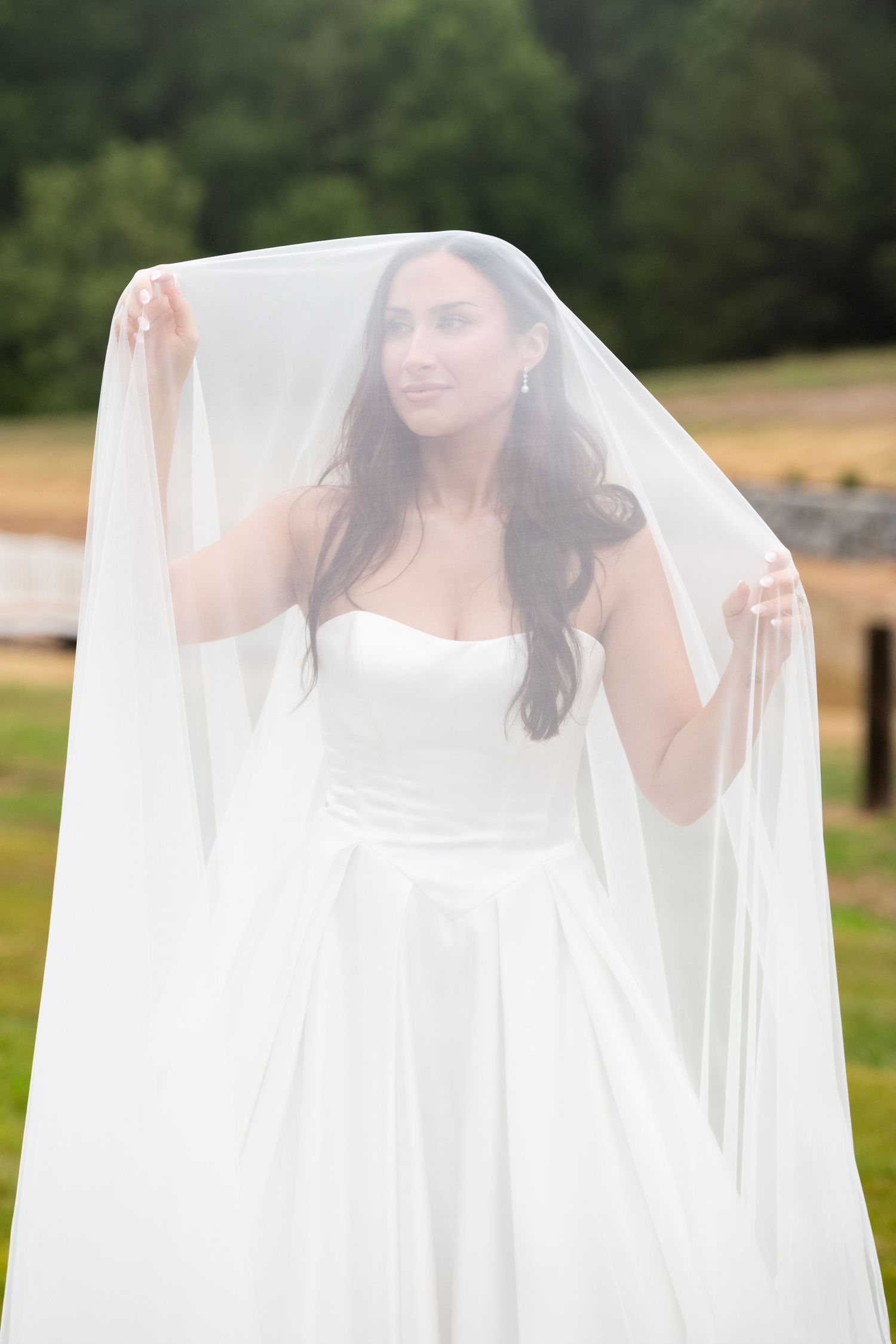 Bride in white strapless gown holding veil overhead outdoors with green trees in background.