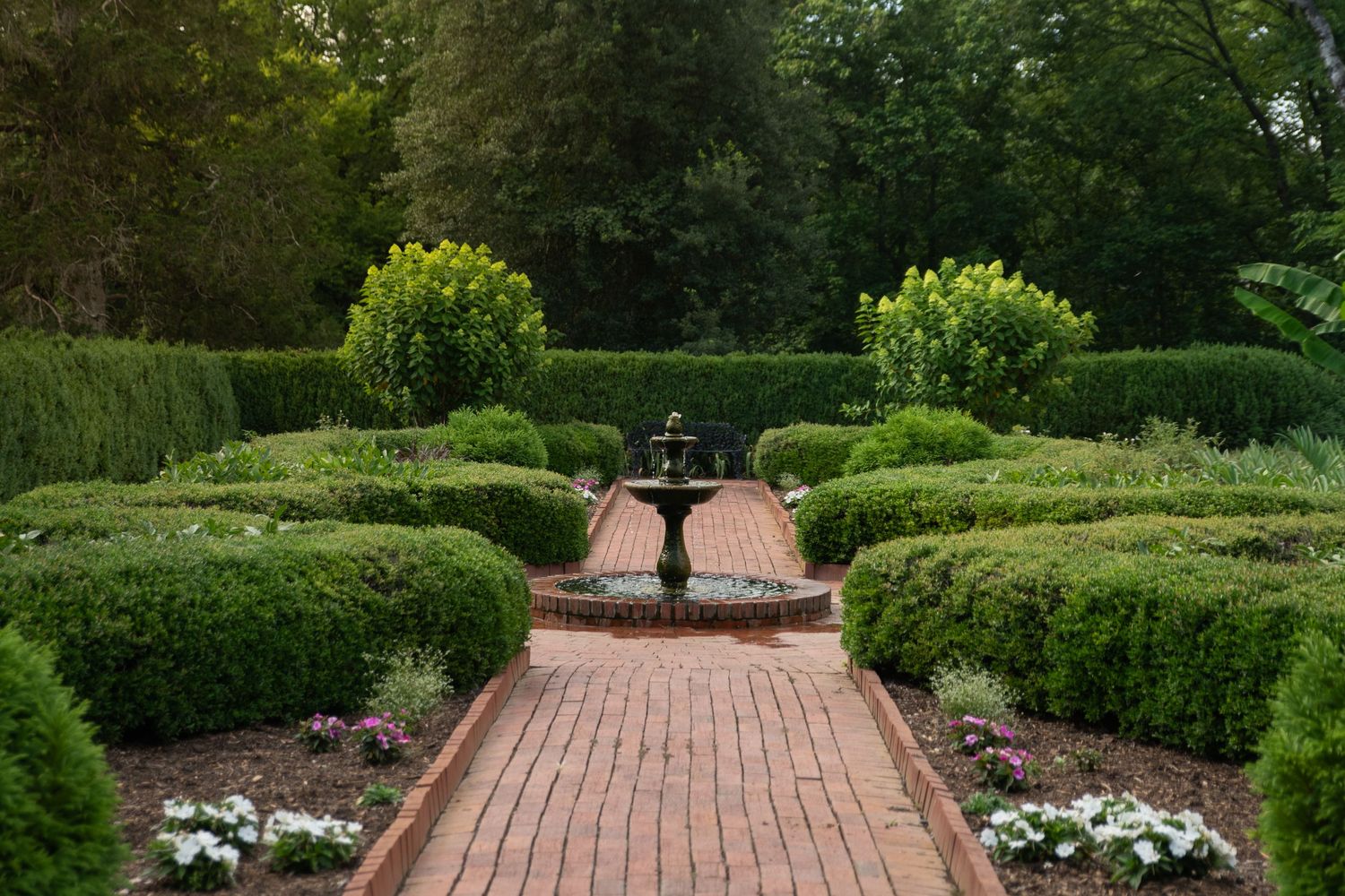 Formal garden with brick pathway leading to ornate fountain, surrounded by neatly trimmed hedges and lush green trees.