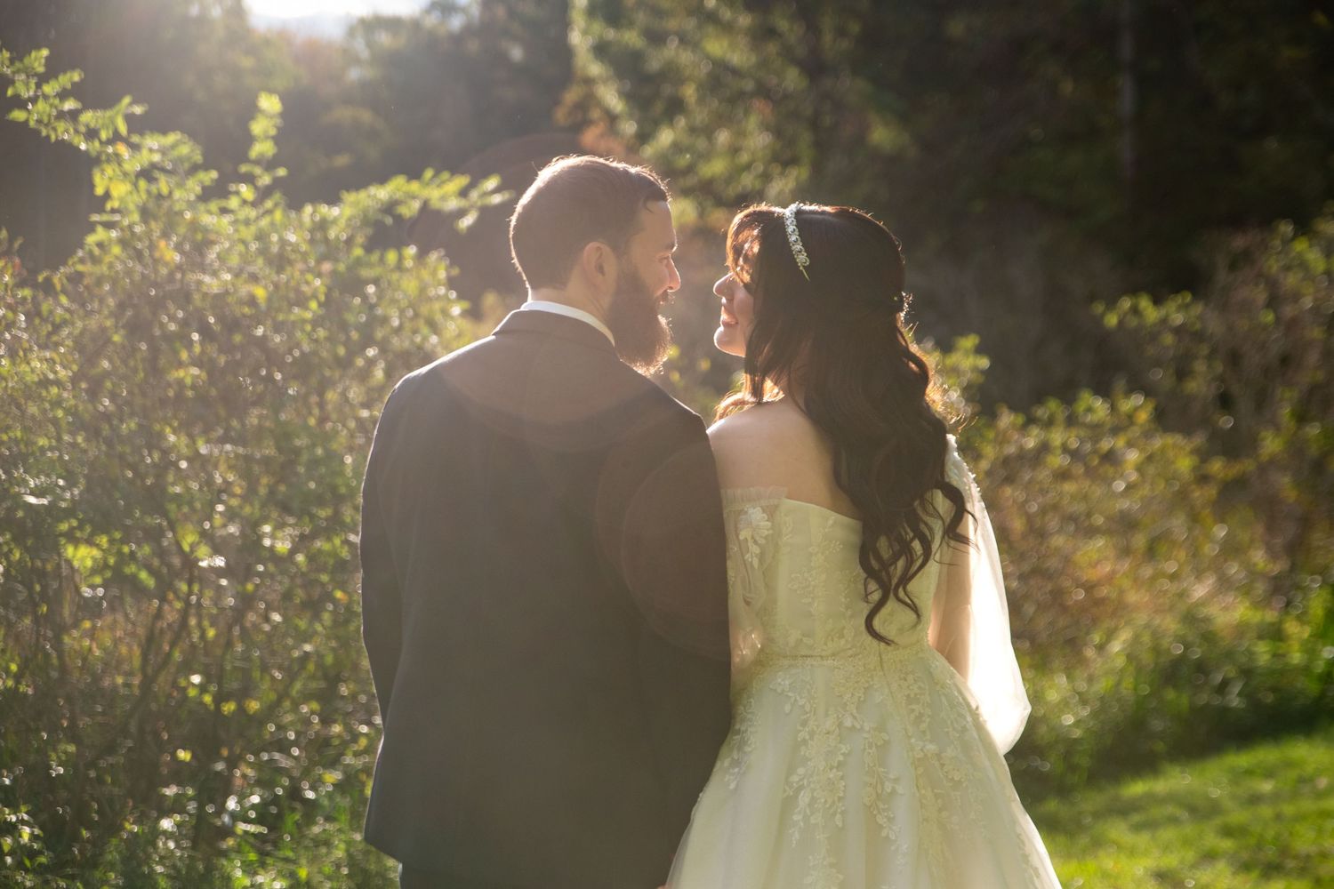 Bride in a lace wedding gown with long dark curls gazes at her groom in a dark suit while standing in a sunlit garden.