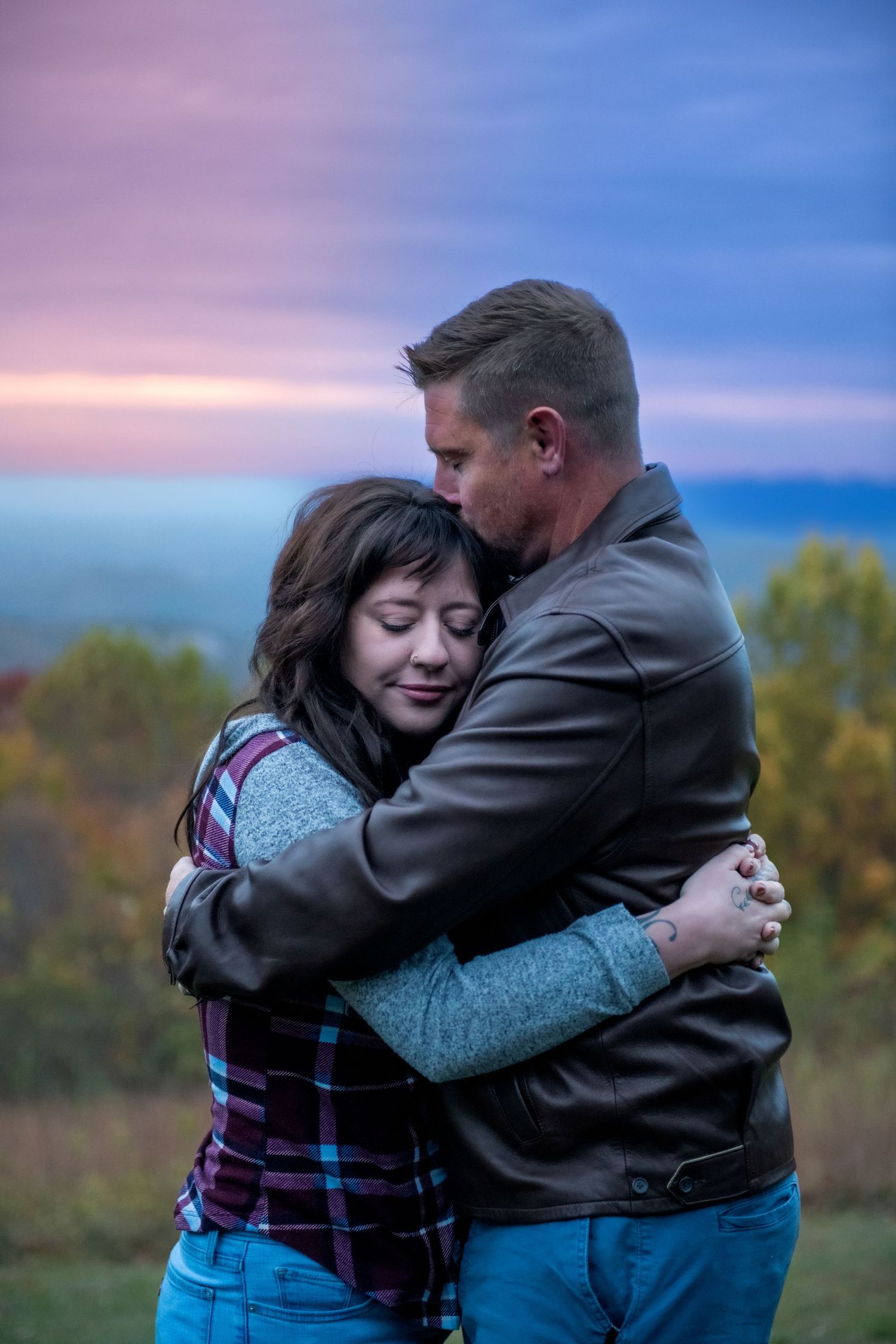 A couple embraces outdoors at sunset with autumn trees in the background during a tender moment.