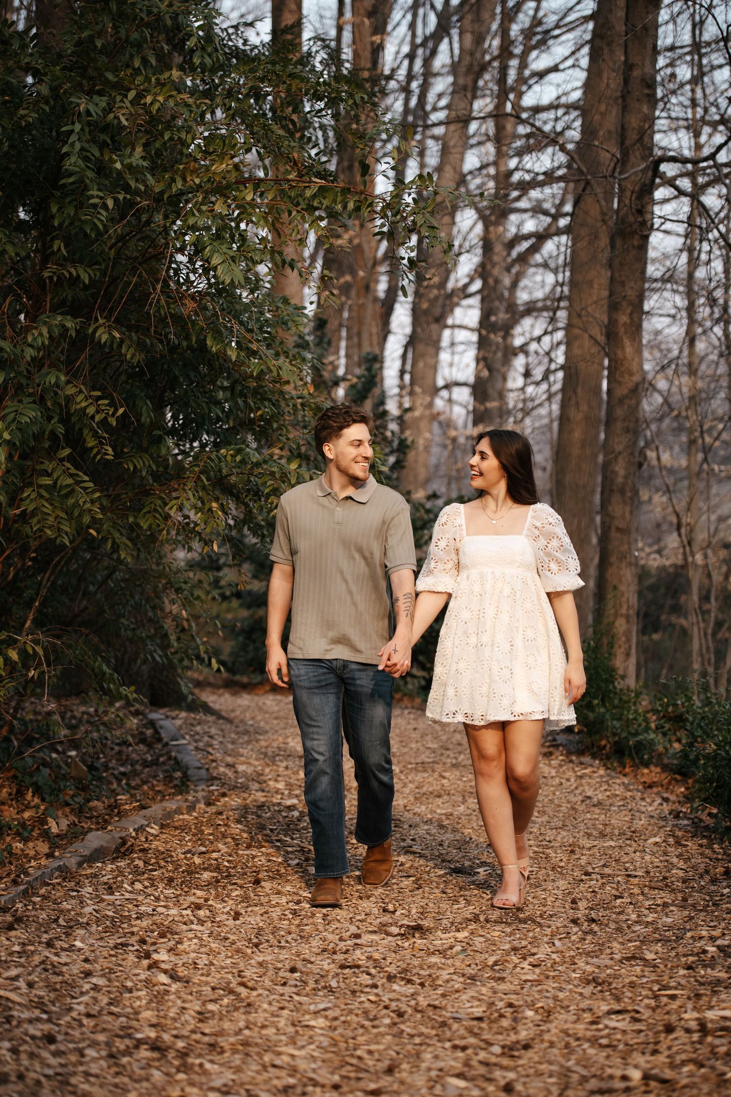 A couple walks hand in hand down a leaf-covered forest path.