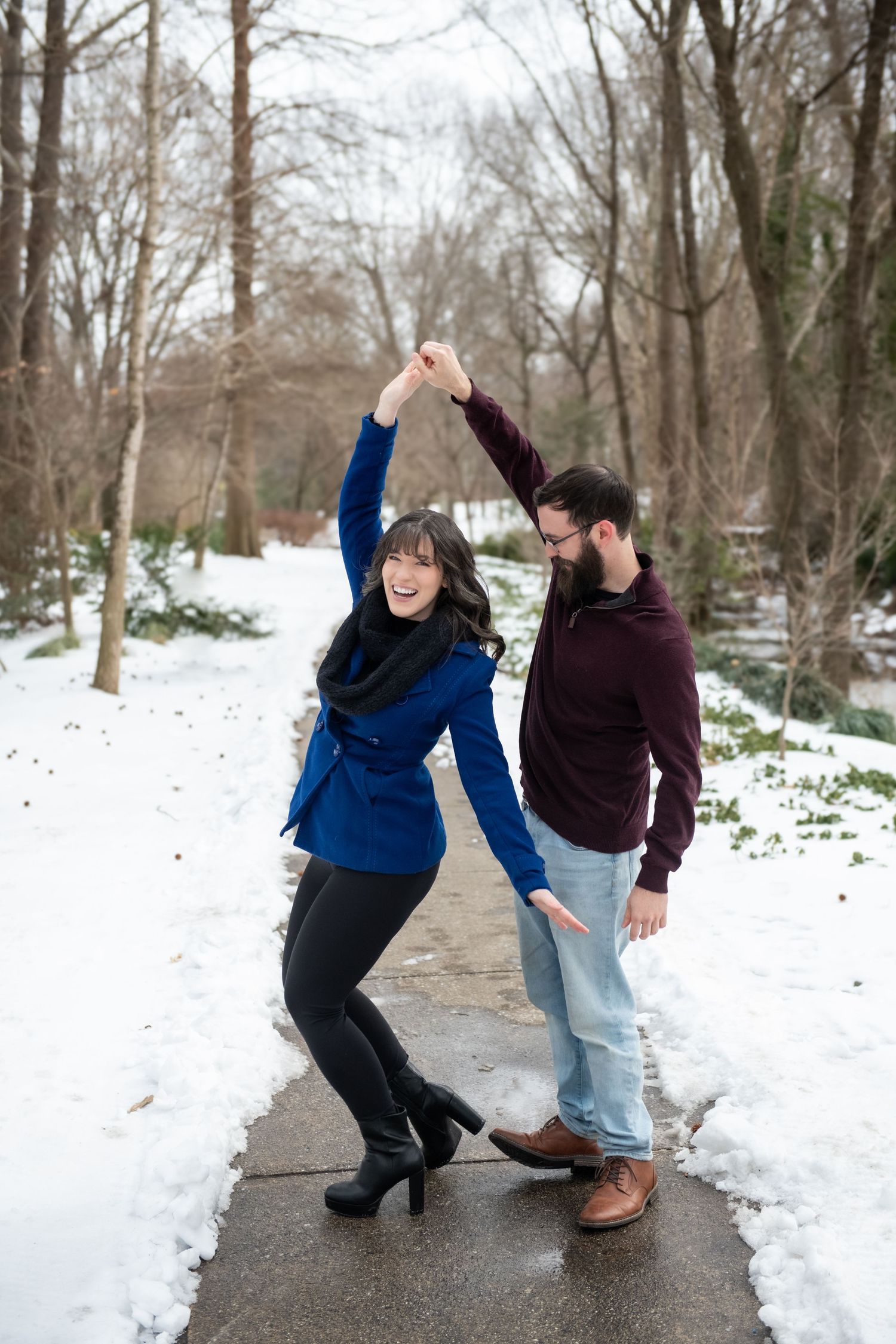A couple dances and plays together on a snow-covered walkway surrounded by bare winter trees.