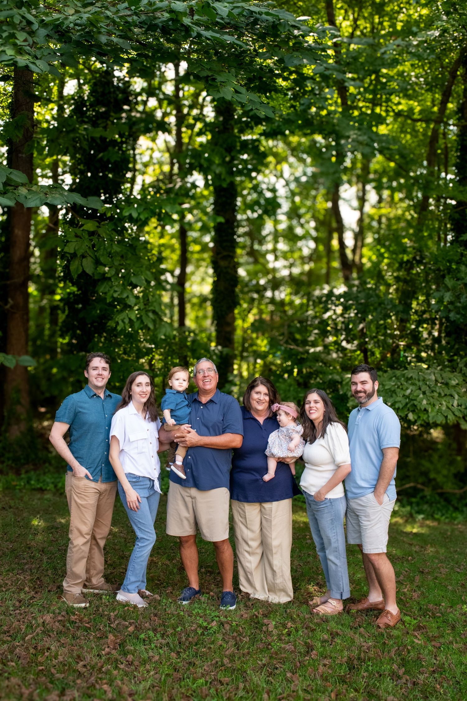 Family group portrait session in a wooded outdoor setting wearing coordinated blue and white casual clothing.