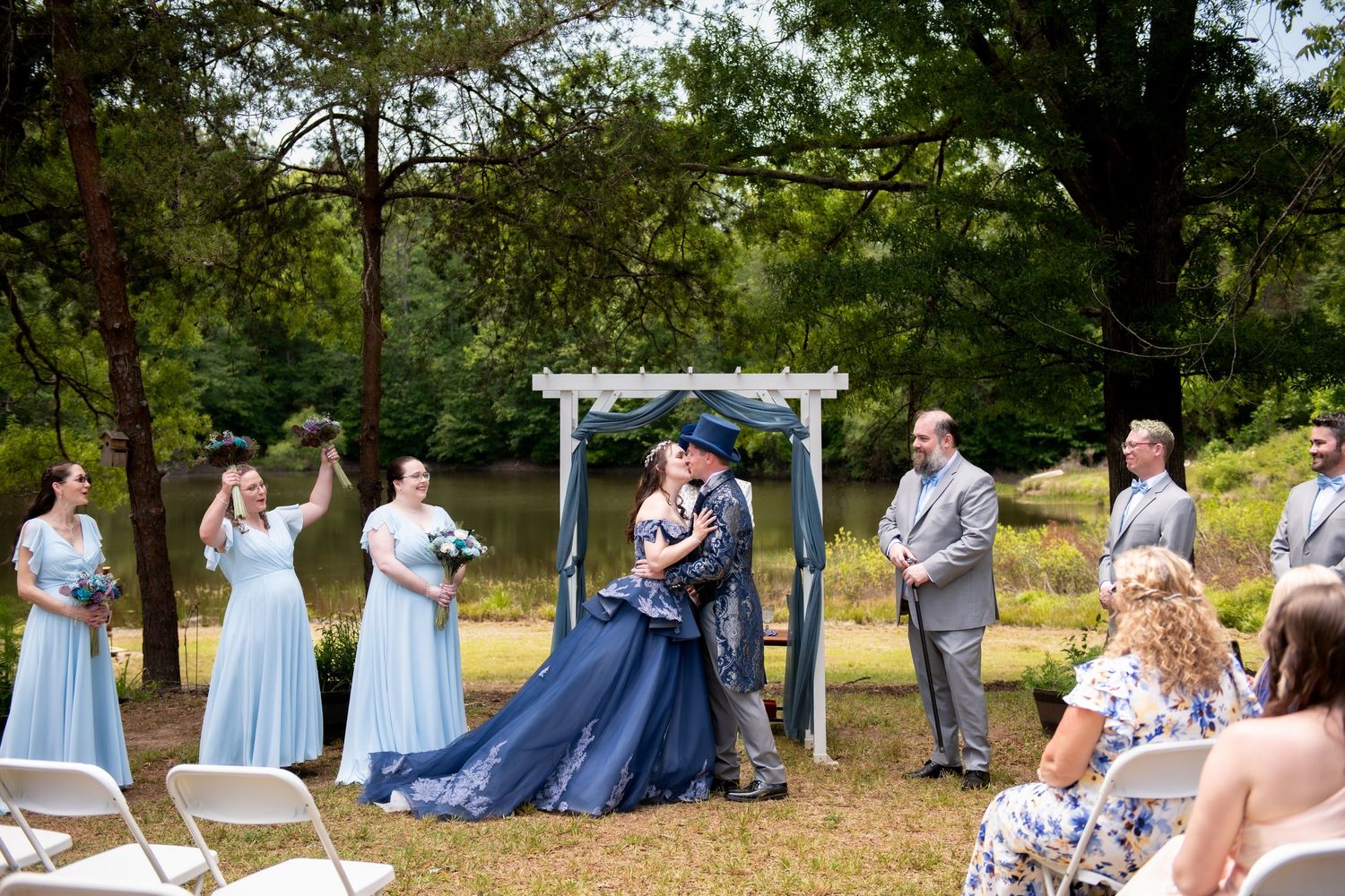 Outdoor wedding ceremony with bride in navy gown, bridesmaids in blue, groomsmen in gray, under white arbor by a pond.