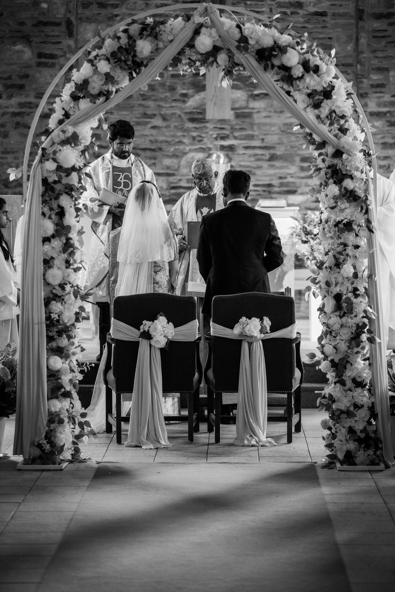 Black and white photo of wedding ceremony under floral arch in brick venue.