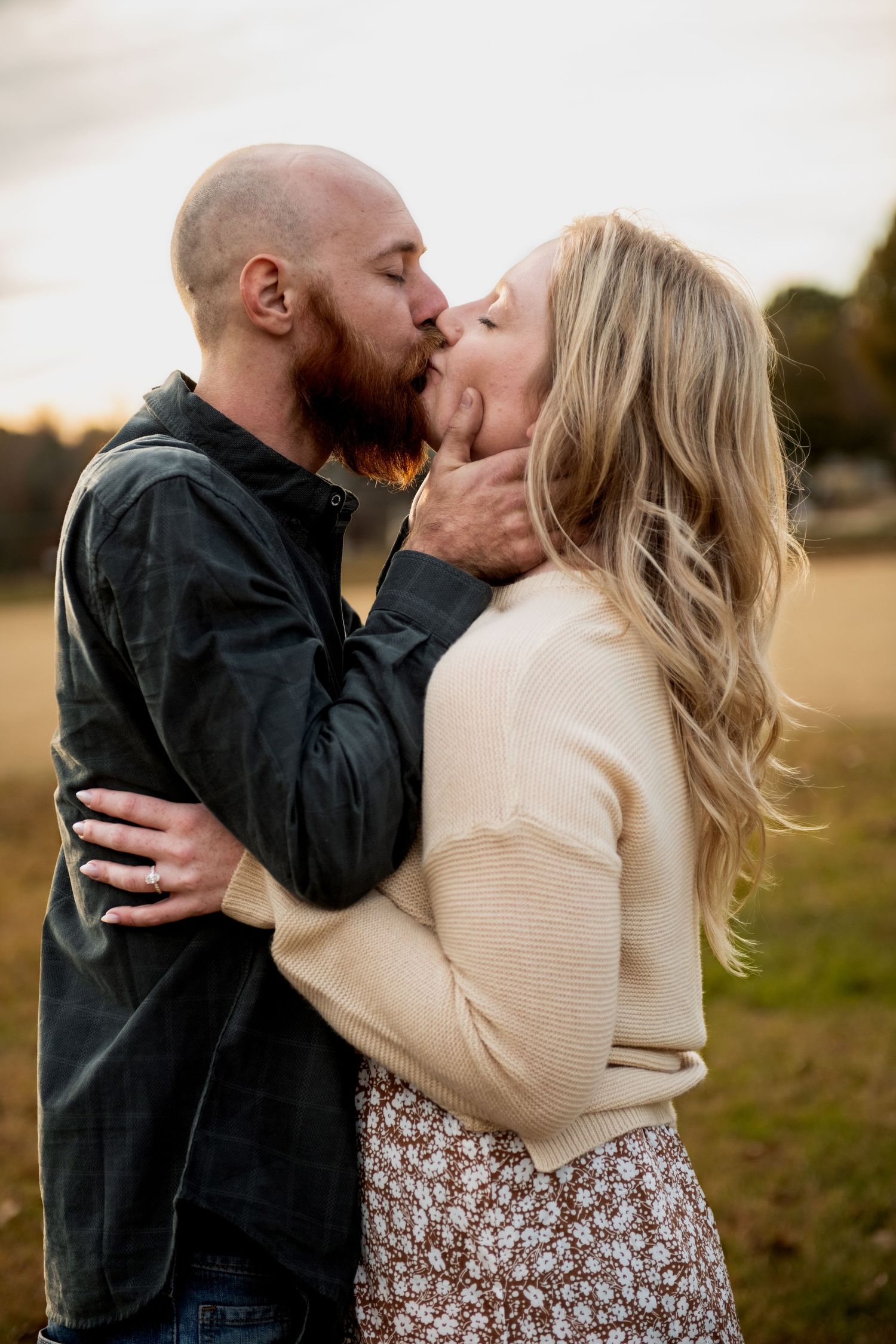 A couple shares a romantic kiss outdoors at sunset, with her wearing a cream sweater and floral skirt.