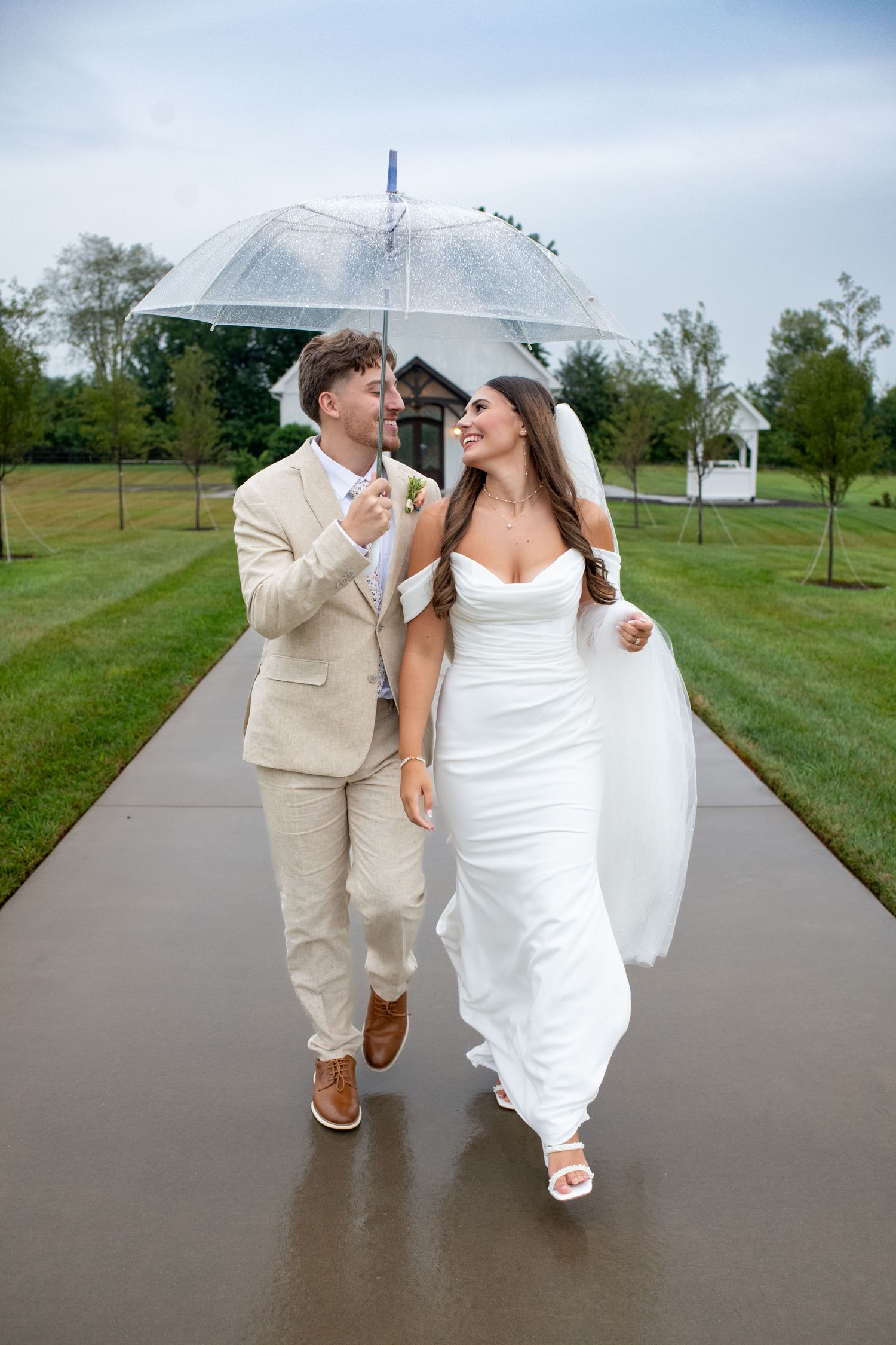 A couple shares a clear umbrella while walking down a wet path at an outdoor wedding venue.