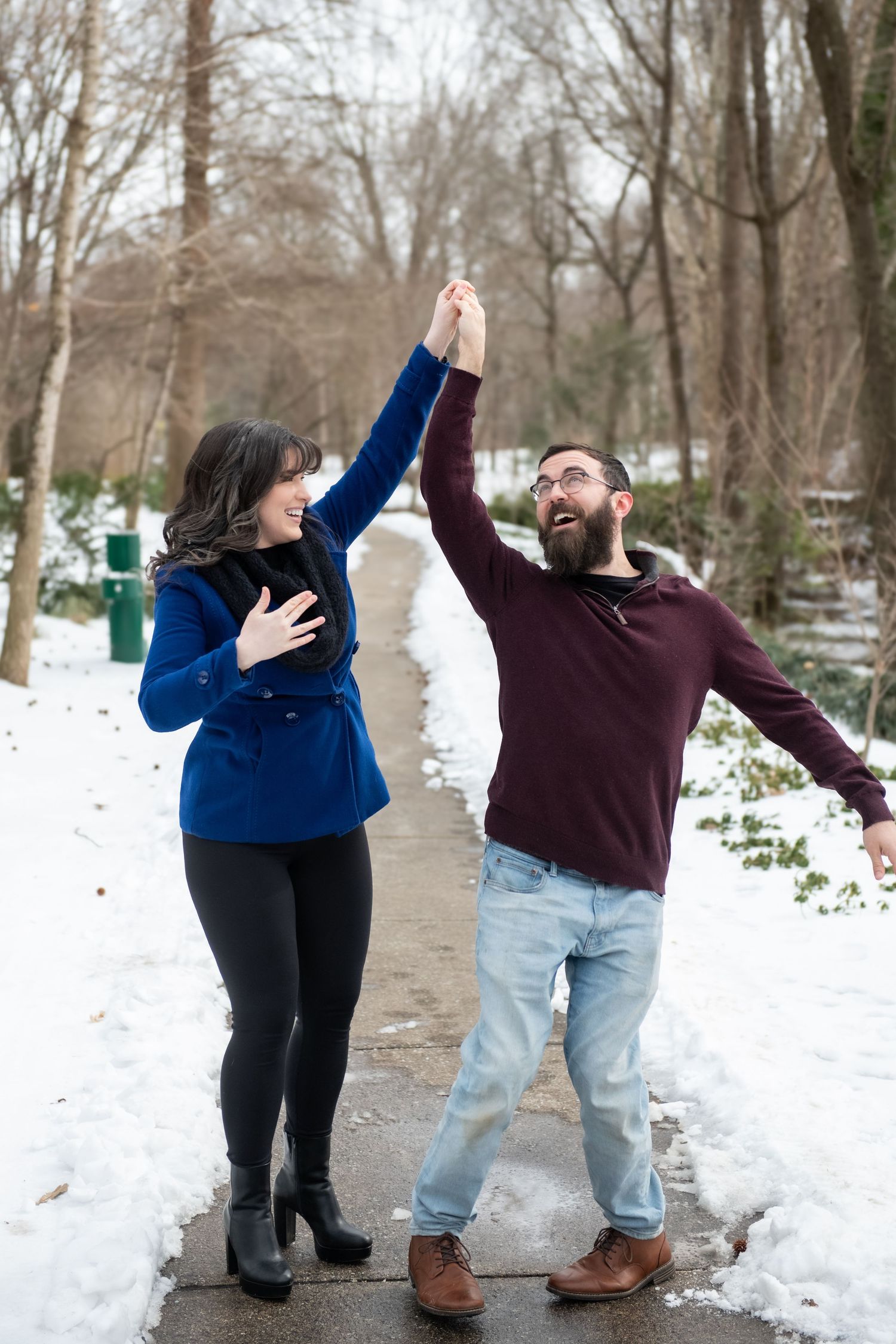 A couple dances playfully together on a snowy path surrounded by winter trees.