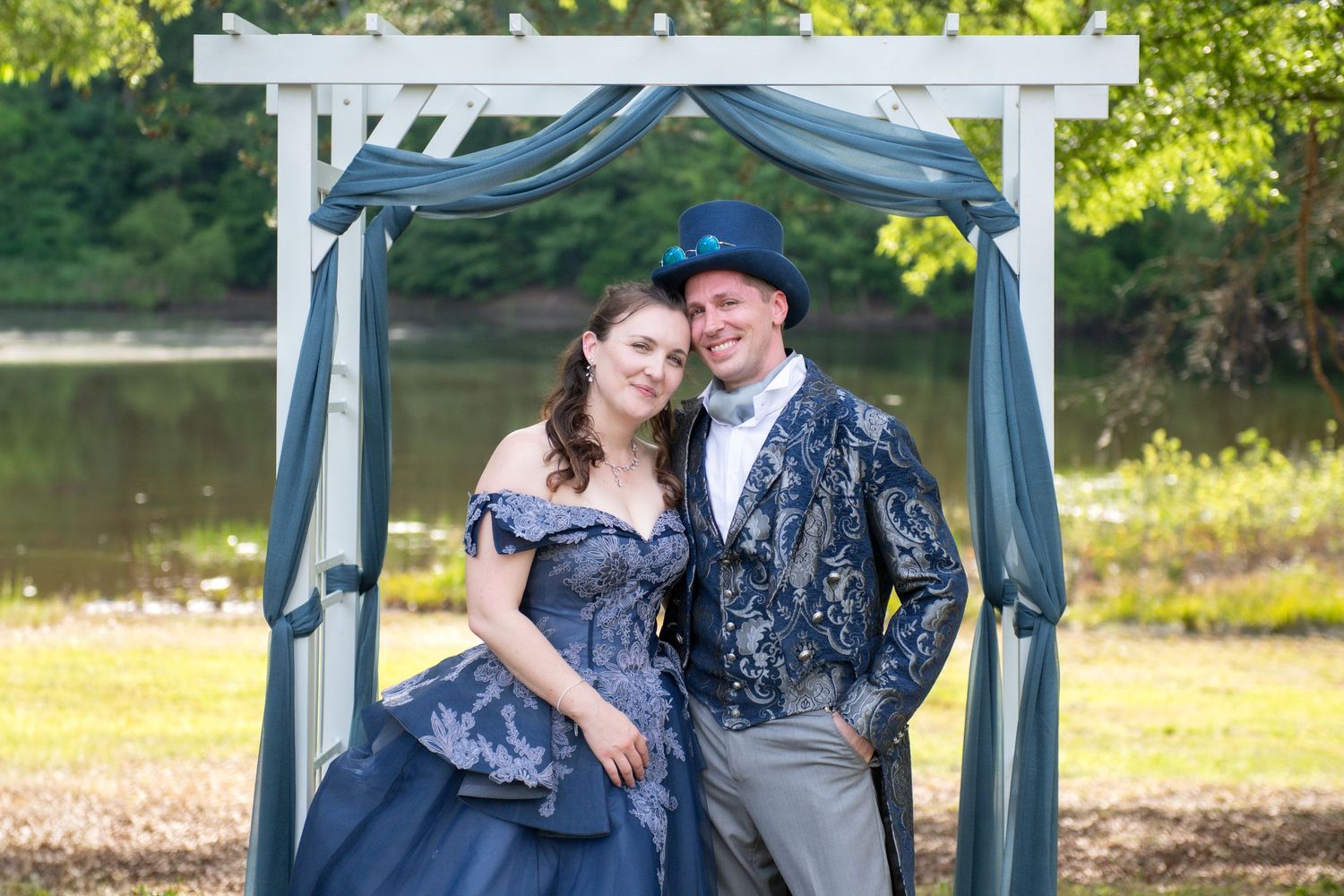 Couple in matching blue Victorian steampunk wedding attire posing under a white arbor draped with teal fabric outdoors.