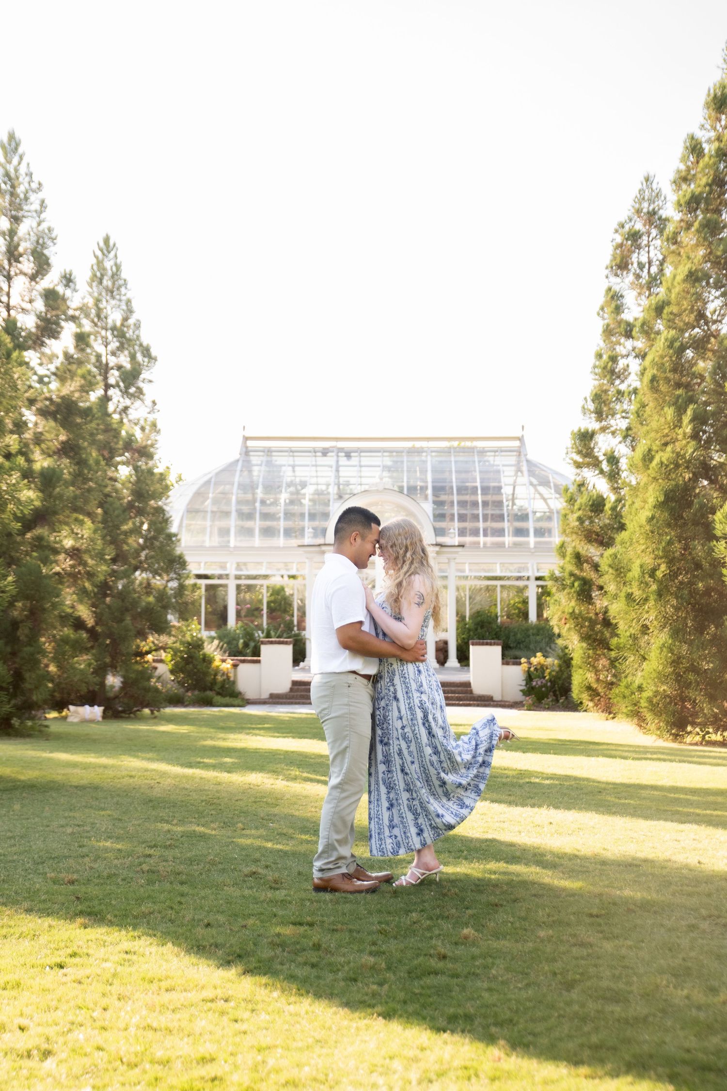 Couple sharing a romantic moment on green lawn before a glass greenhouse, surrounded by tall trees.