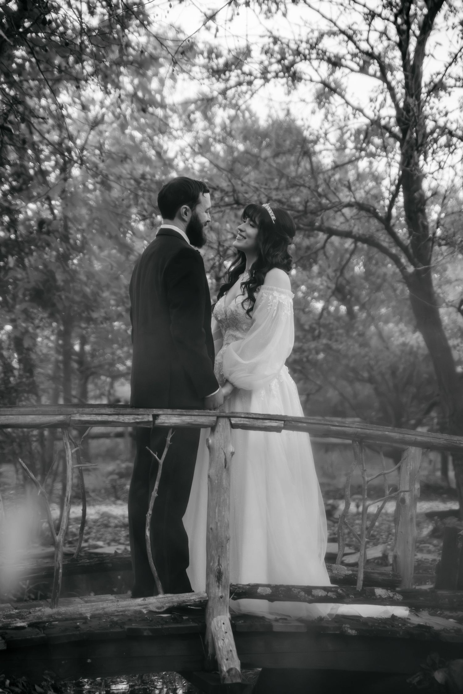 Black and white photo of a bride and groom sharing a loving glance on a rustic wooden bridge surrounded by trees.