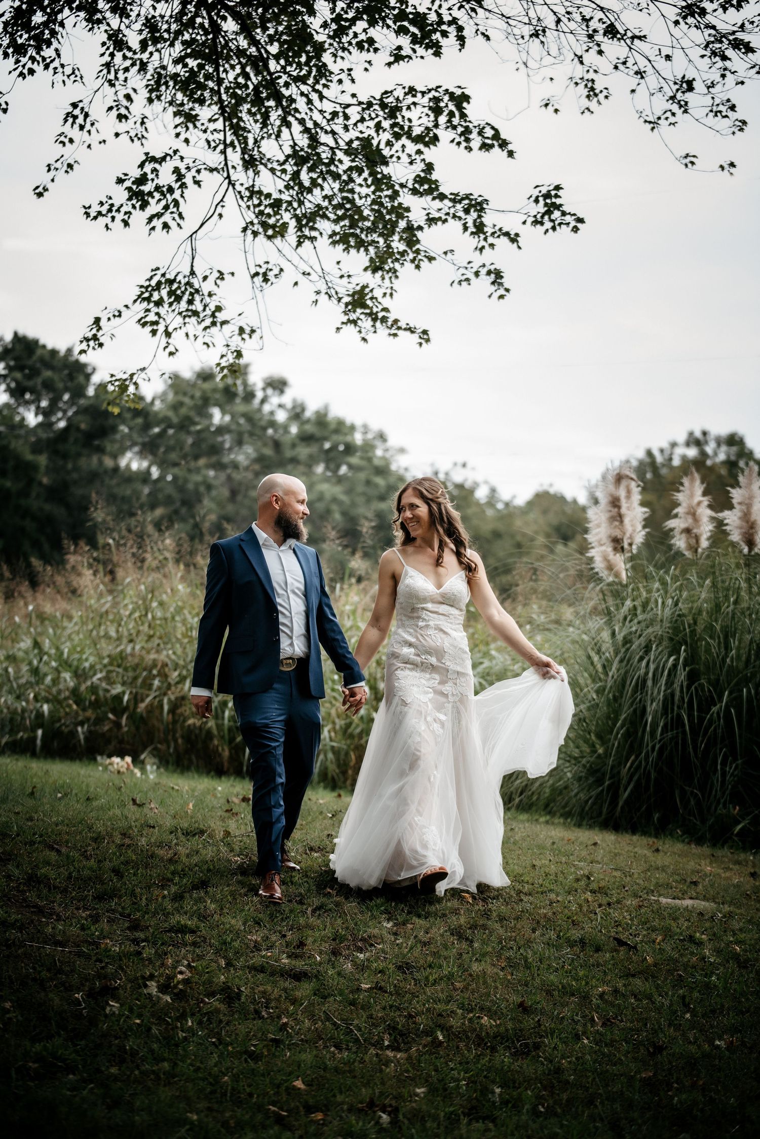A romantic wedding couple walks hand in hand through a lush garden setting with overhanging tree branches.