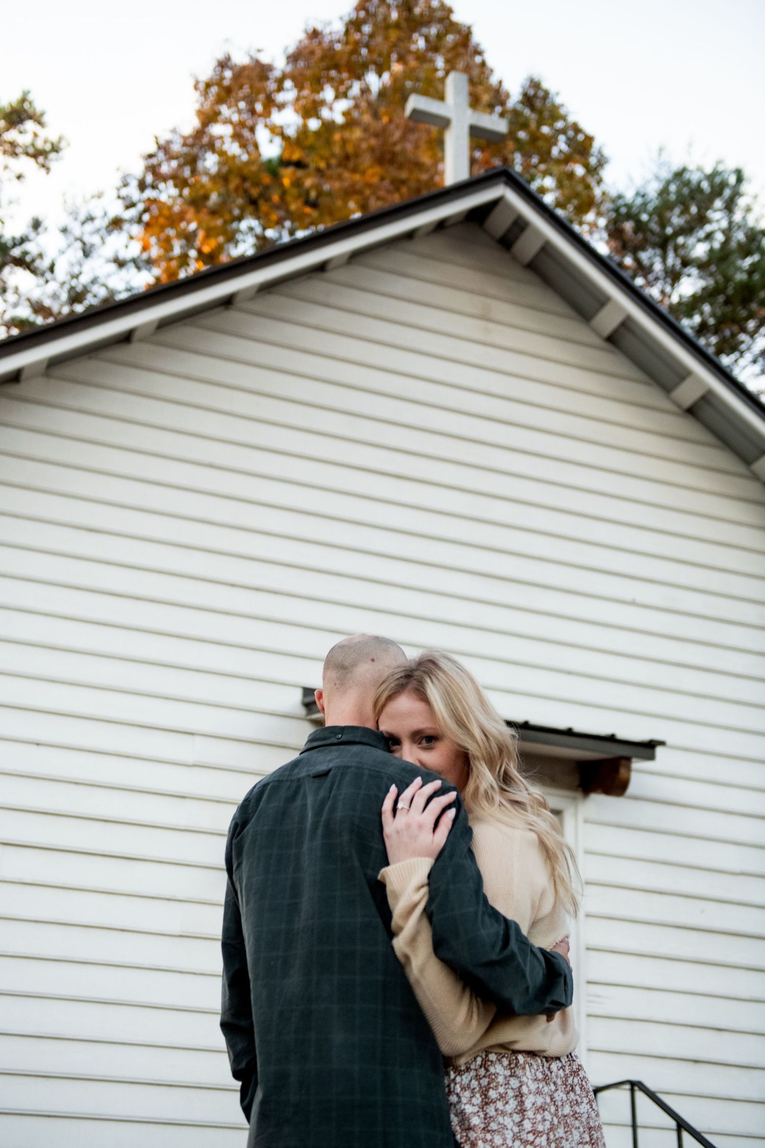 A couple embraces outside a white building with autumn trees in the background.