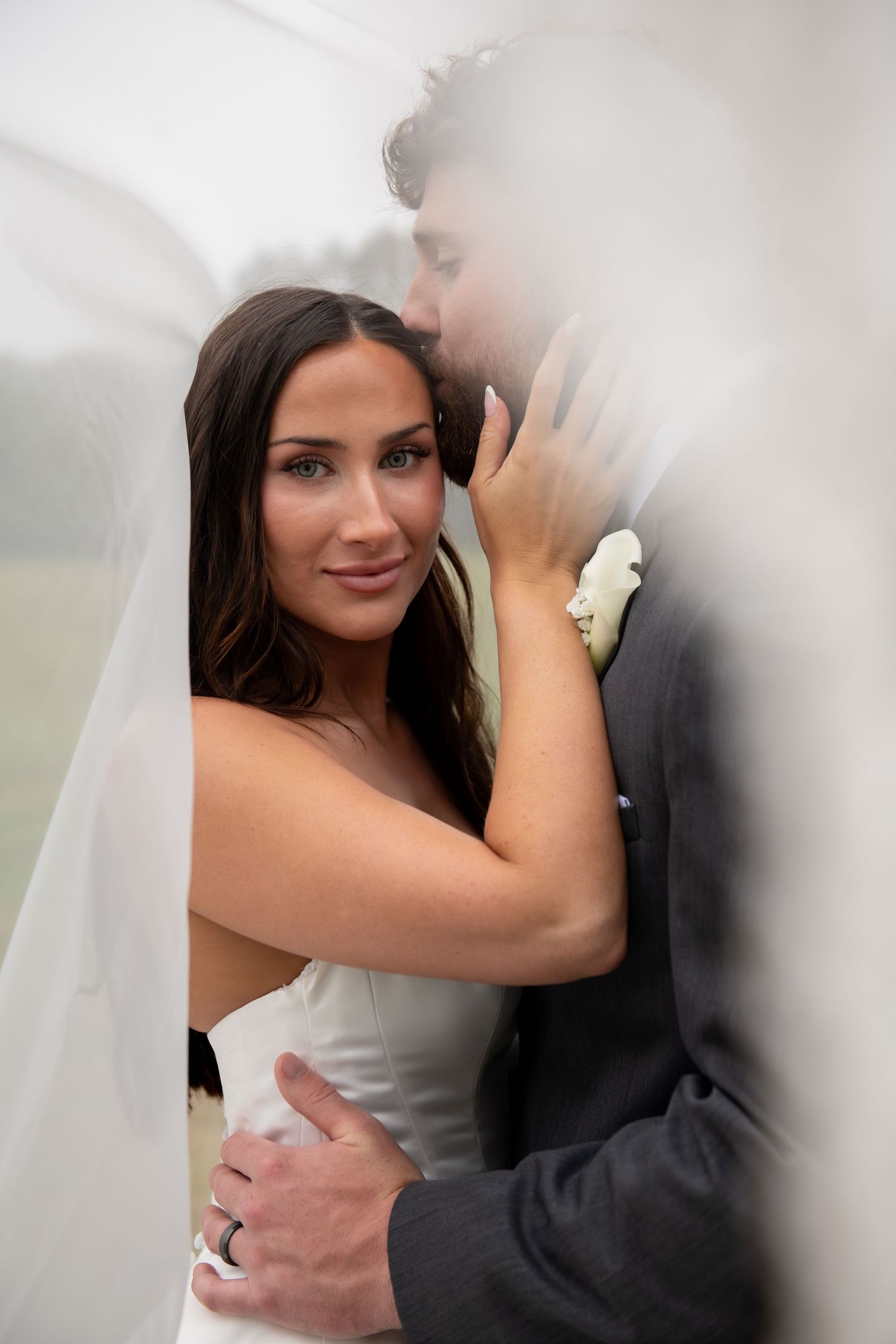 A bride smiles softly as the groom kisses her cheek, both embracing under a flowing white veil.