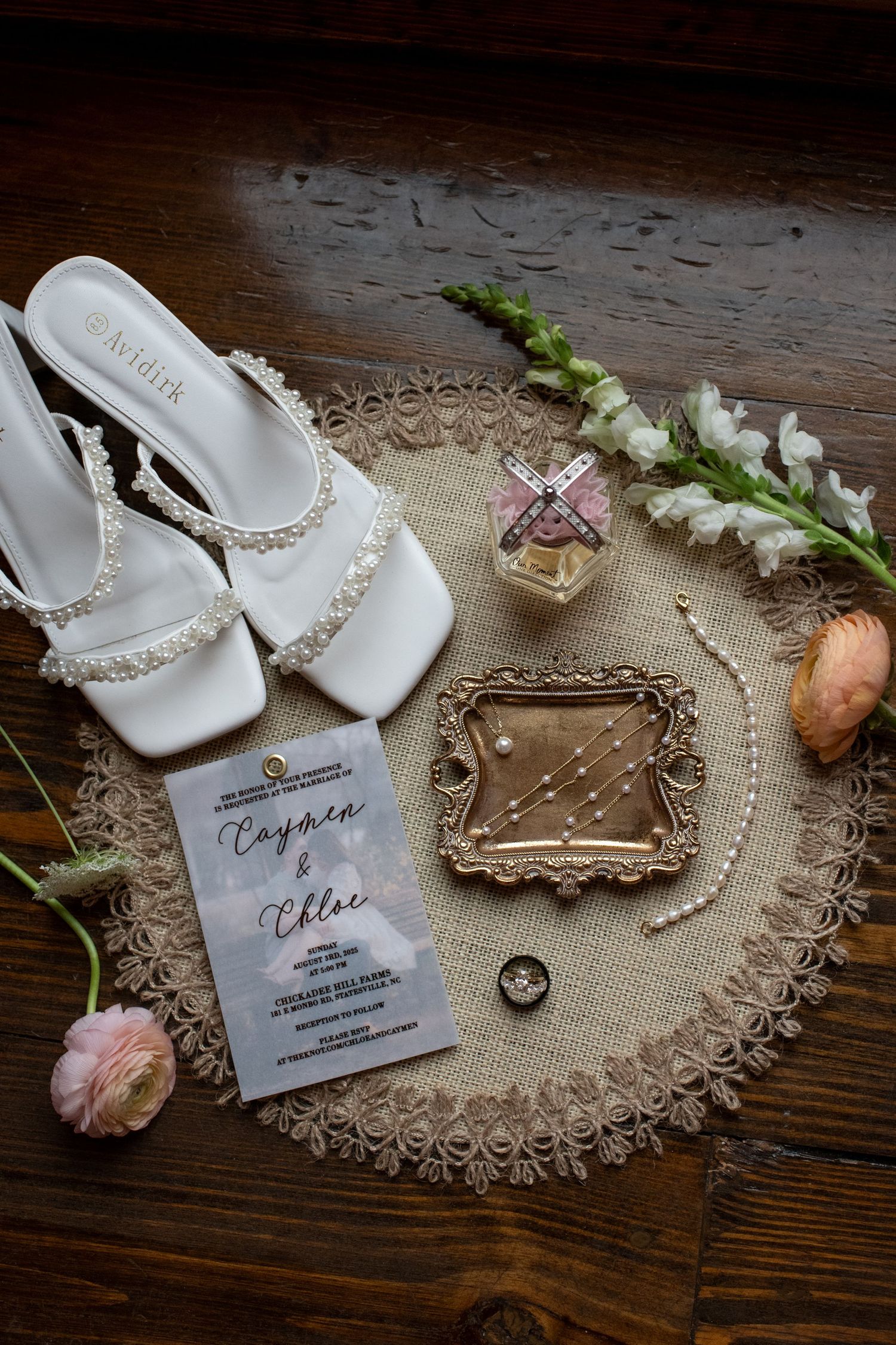 Wedding accessories including white sandals, invitation, and flowers arranged on rustic wood.