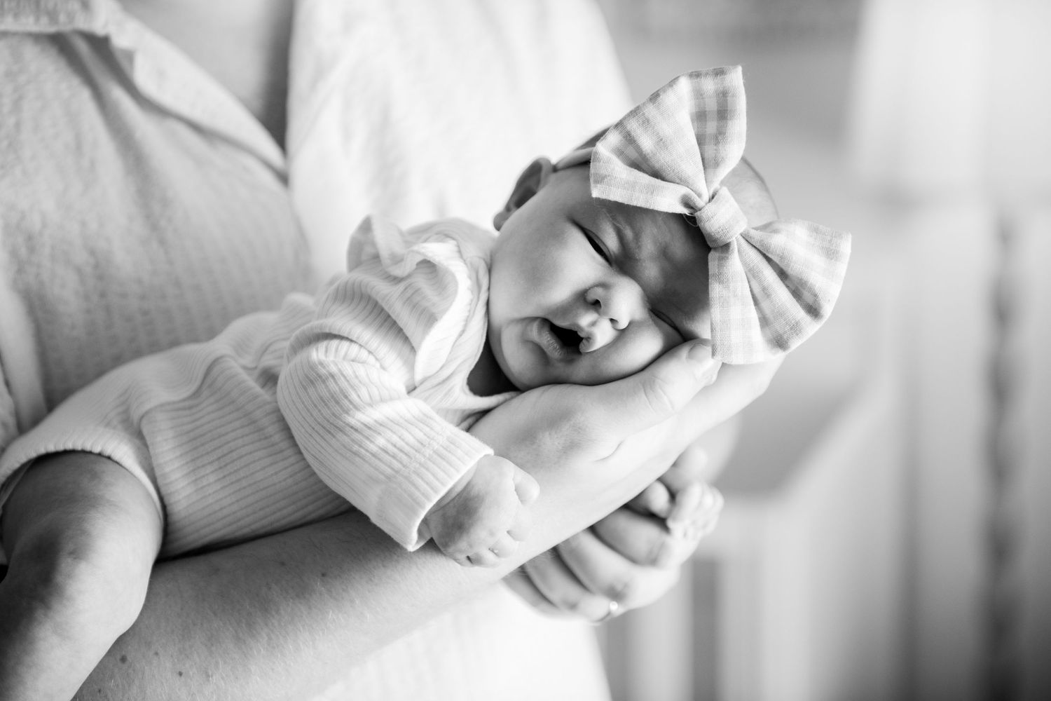 Black and white photo of a sleeping newborn baby in a bow headband cradled in an adult's arms.