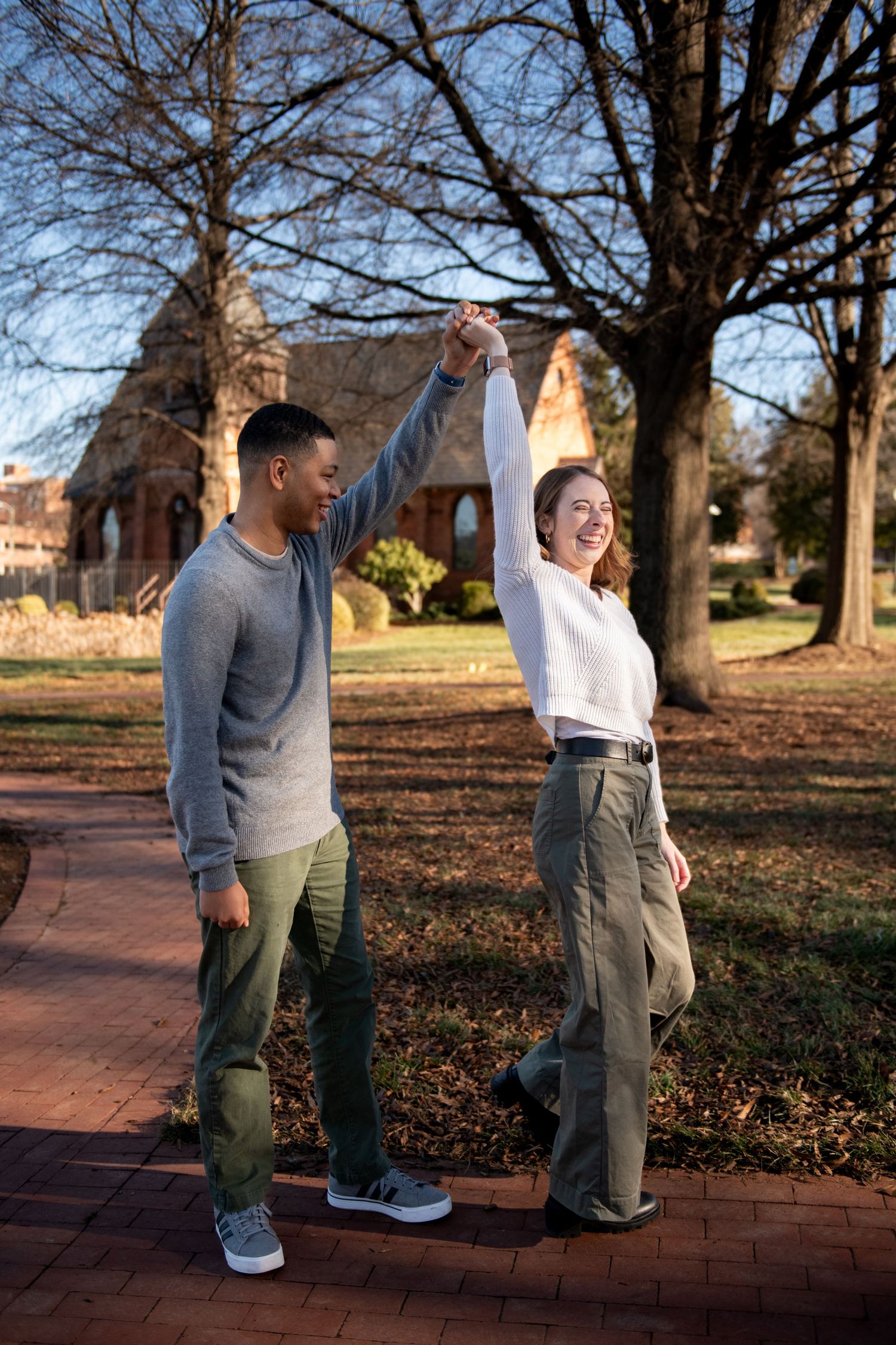 A joyful couple holds hands and raises their arms while standing on a brick path in a park during autumn.