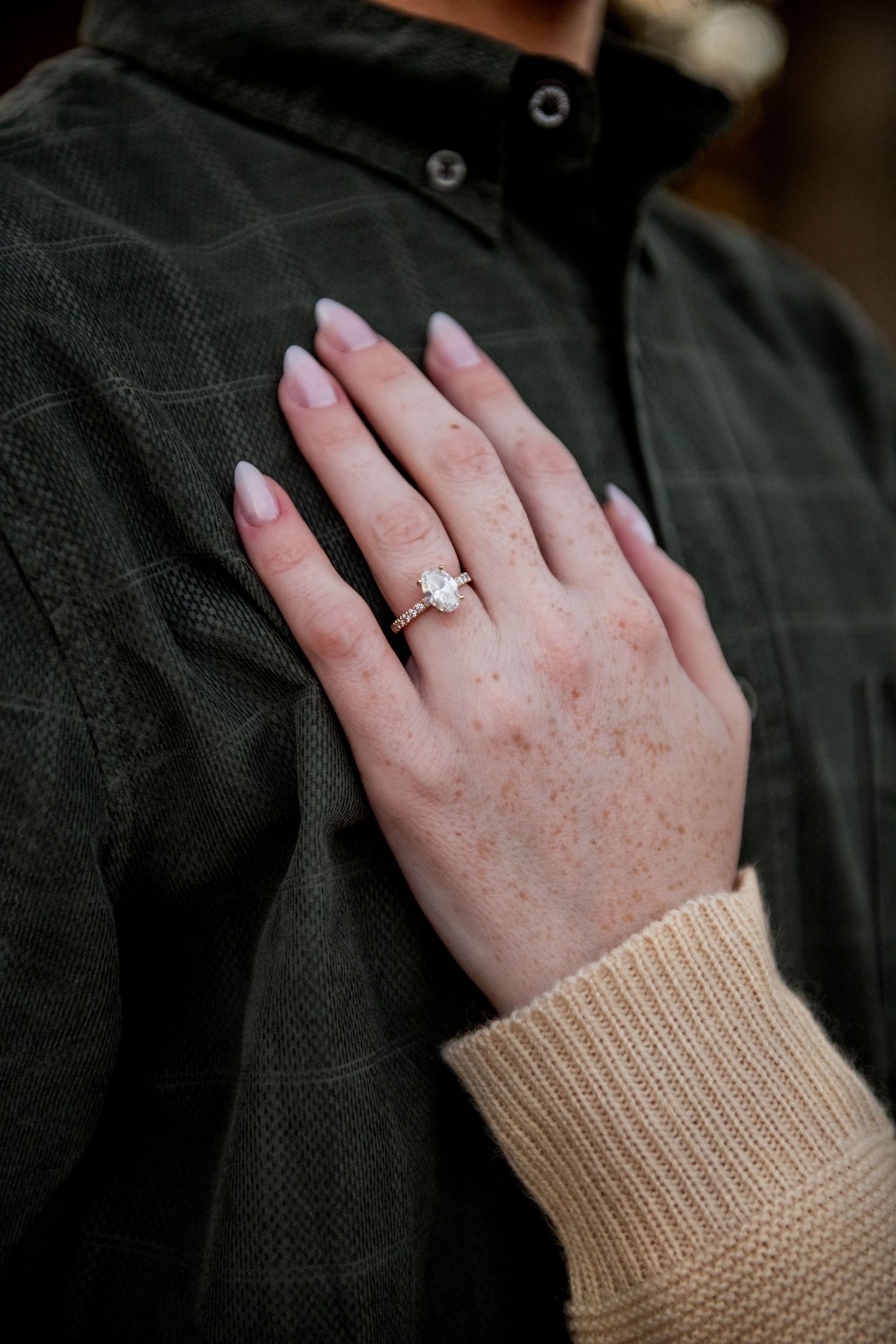 Close-up shot of a hand wearing a knit beige sweater sleeve and diamond engagement ring against a dark background.