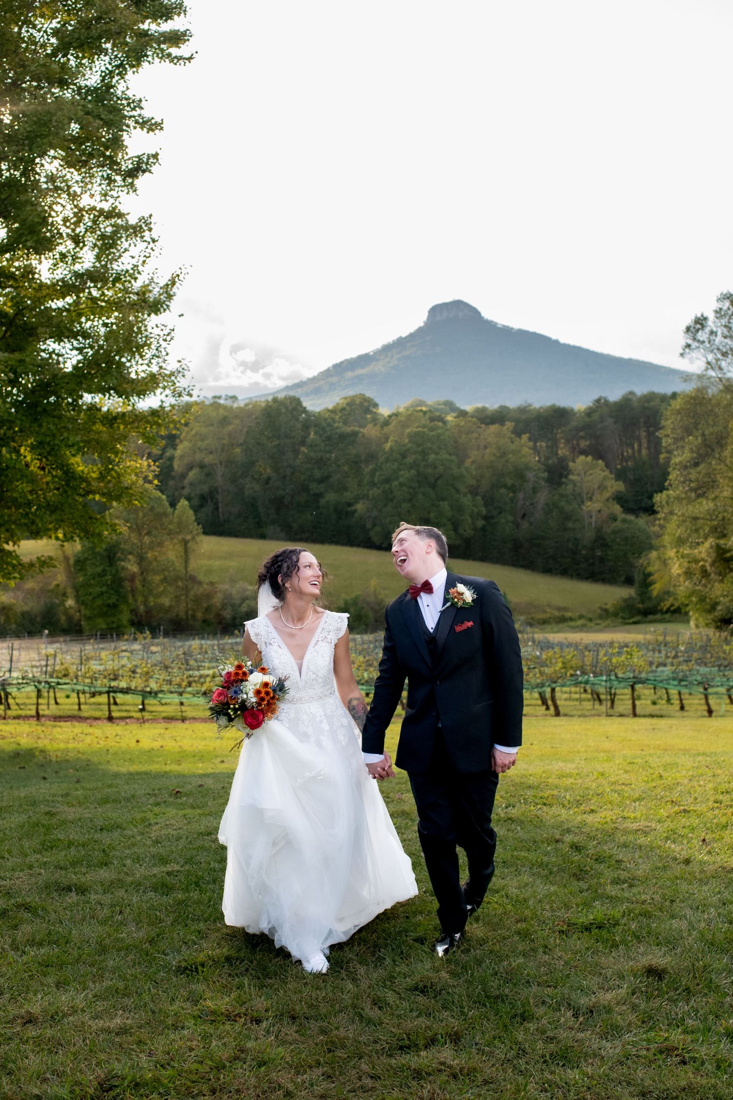 A couple poses in their wedding attire against a scenic mountain vineyard backdrop at sunset.