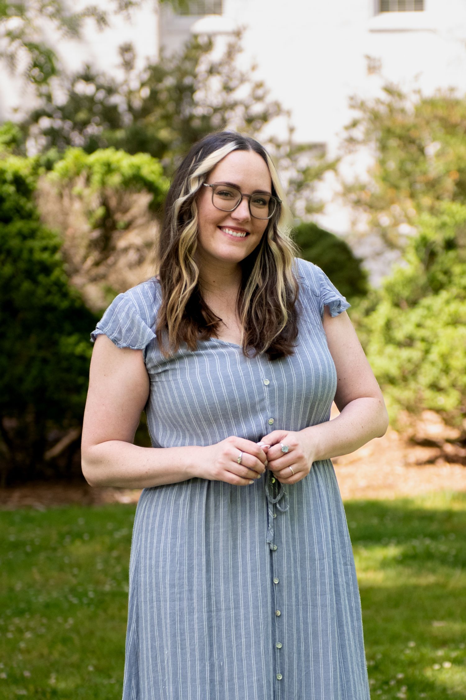 Smiling woman with glasses in a blue striped dress standing outdoors in a green garden setting.