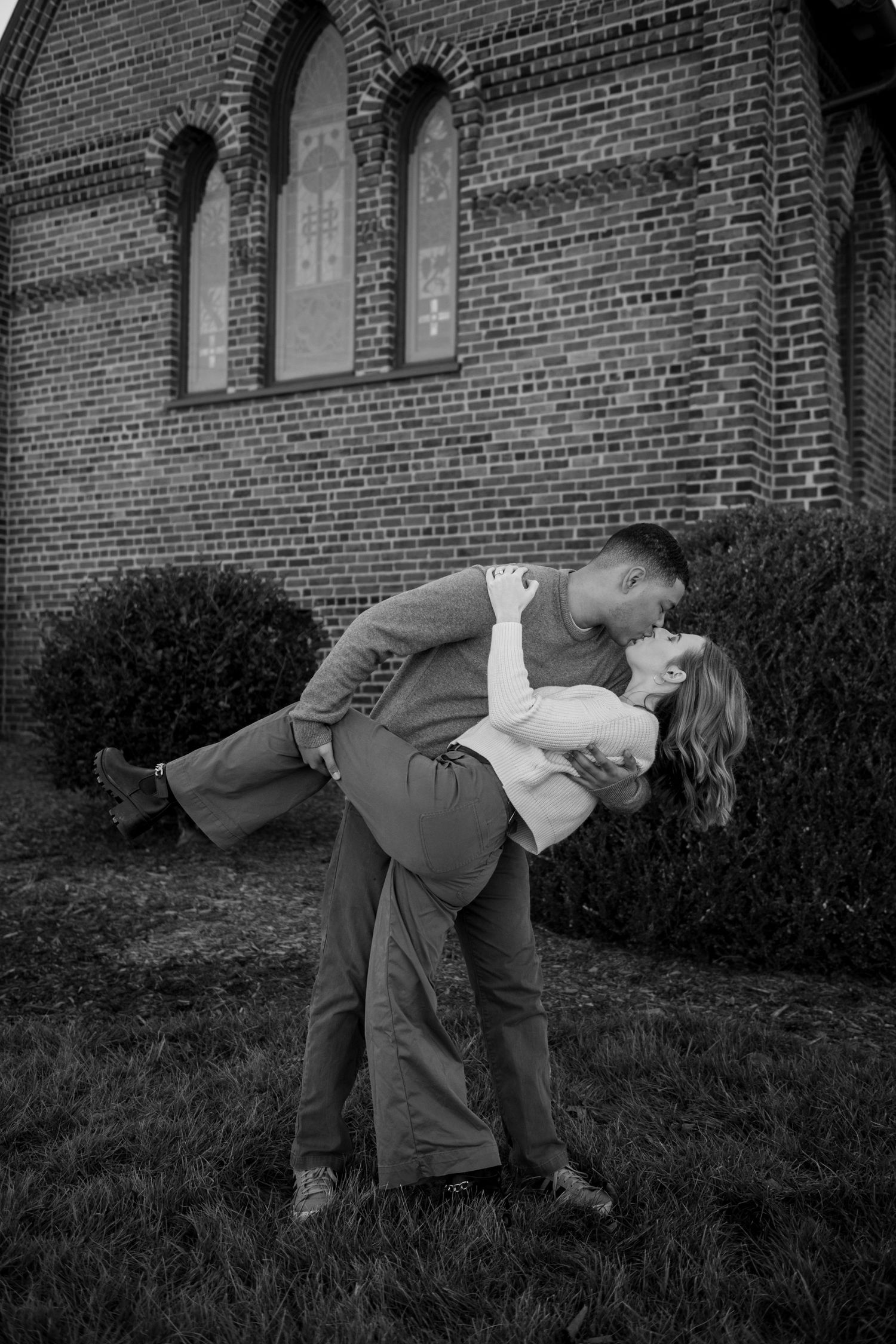 A romantic couple embraces in a dip pose outside a brick church with arched windows.