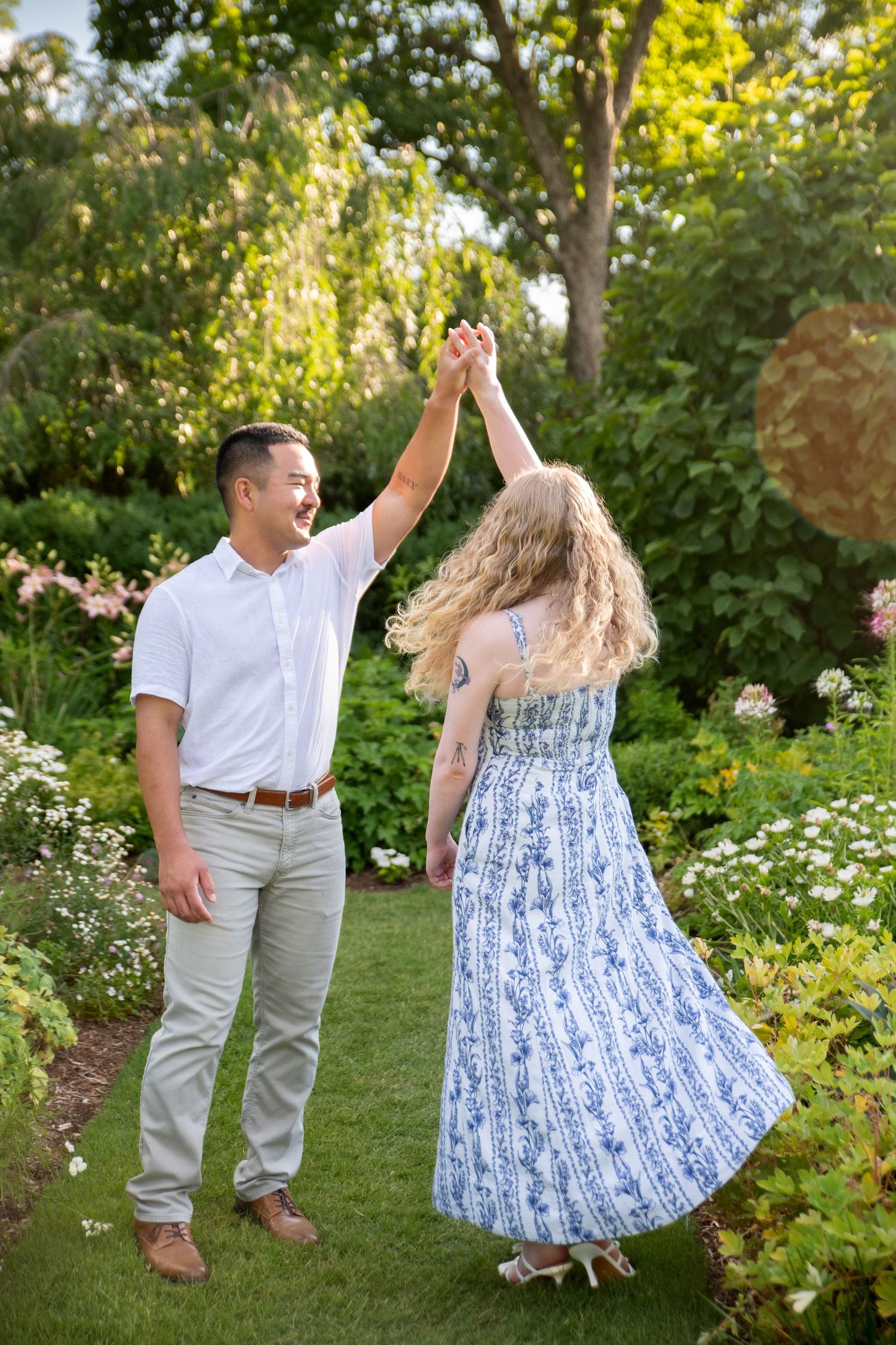 A couple dancing joyfully in a lush garden, man in white shirt twirling woman in blue floral dress.