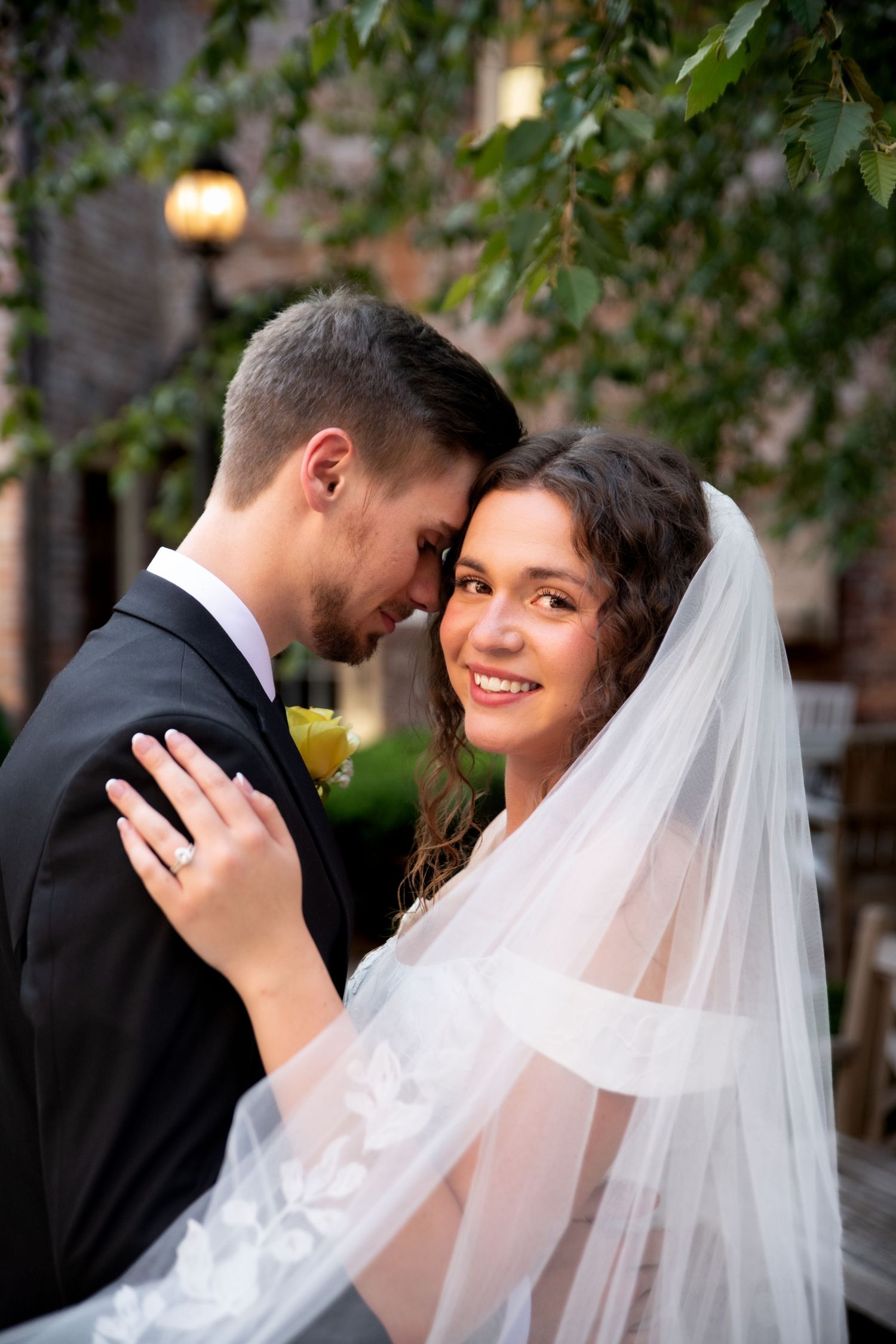 Bride in white veil smiles as groom in black tuxedo leans in close, sharing a romantic moment outdoors.
