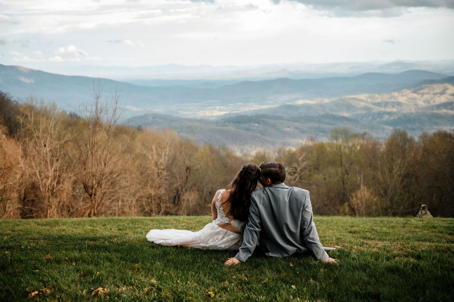 Bride and groom sit together on a grassy hilltop overlooking a stunning mountain valley landscape.