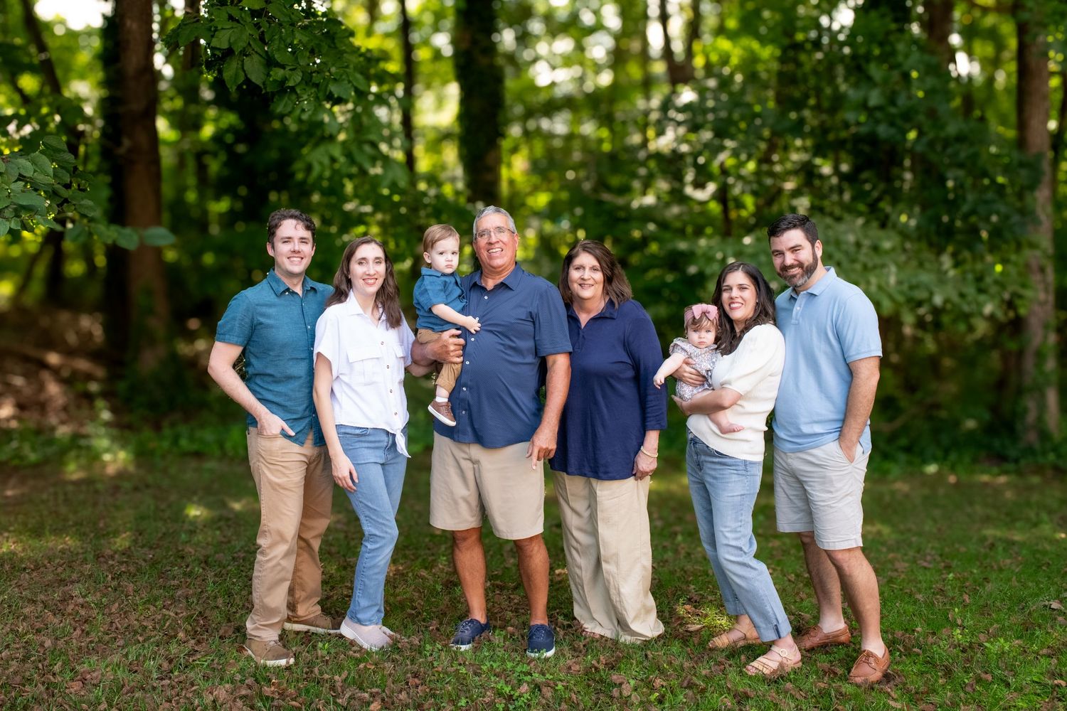 Family of seven poses outdoors in lush green park setting, including two babies, for a professional family portrait.