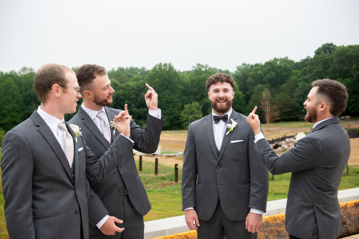 Groomsmen in gray suits pointing at the groom outdoors at a wedding venue with green trees in background.