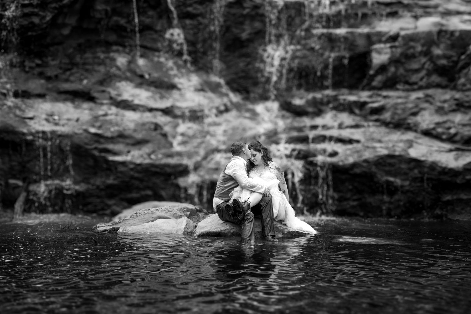 A black and white photo of a couple sitting on rocks by a cascading waterfall.