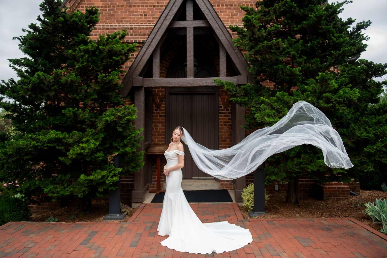 Bride in white gown with flowing veil outside brick chapel surrounded by evergreen trees.