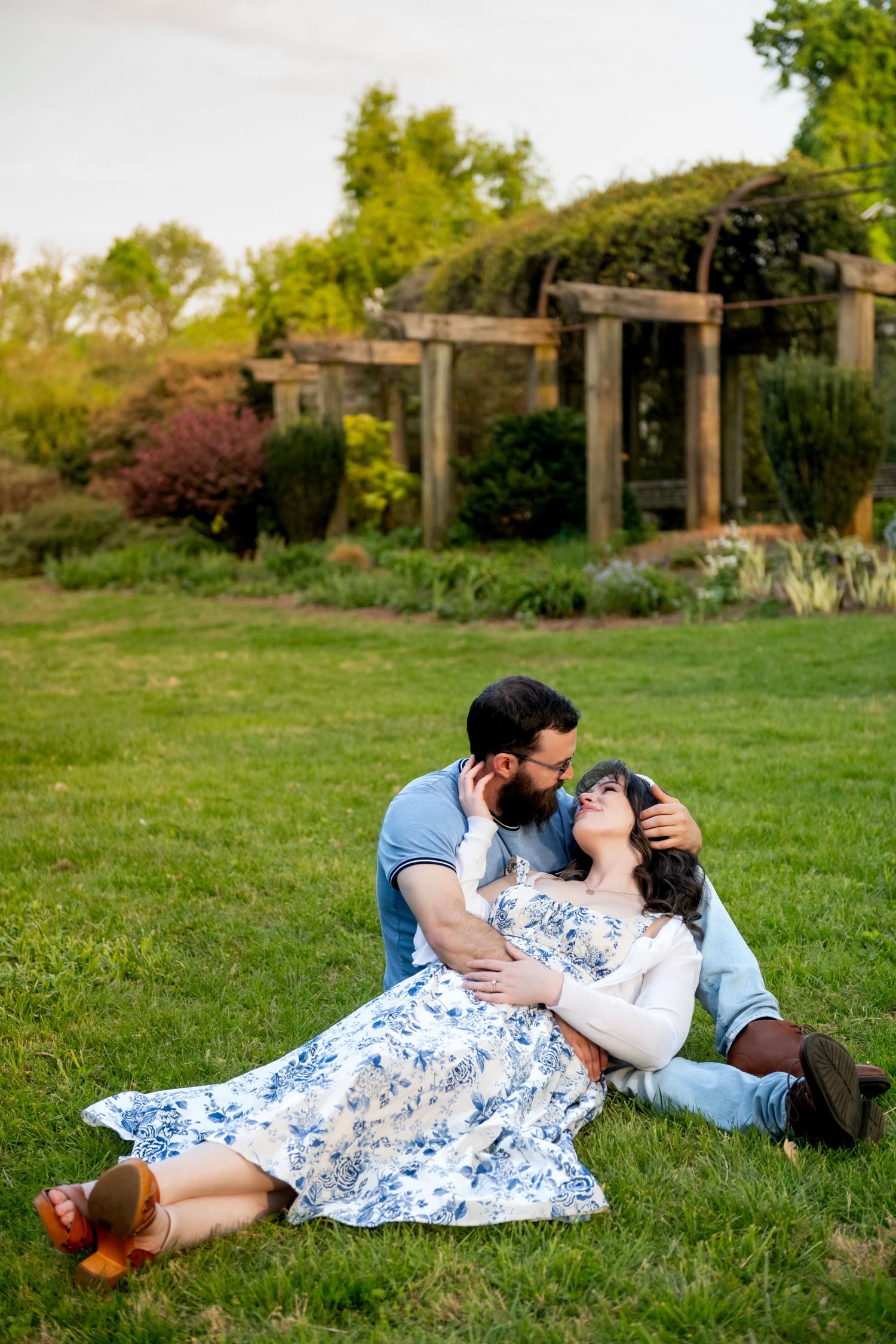Couple sitting on grass in garden, woman in floral dress leaning back into mans arms at golden hour.