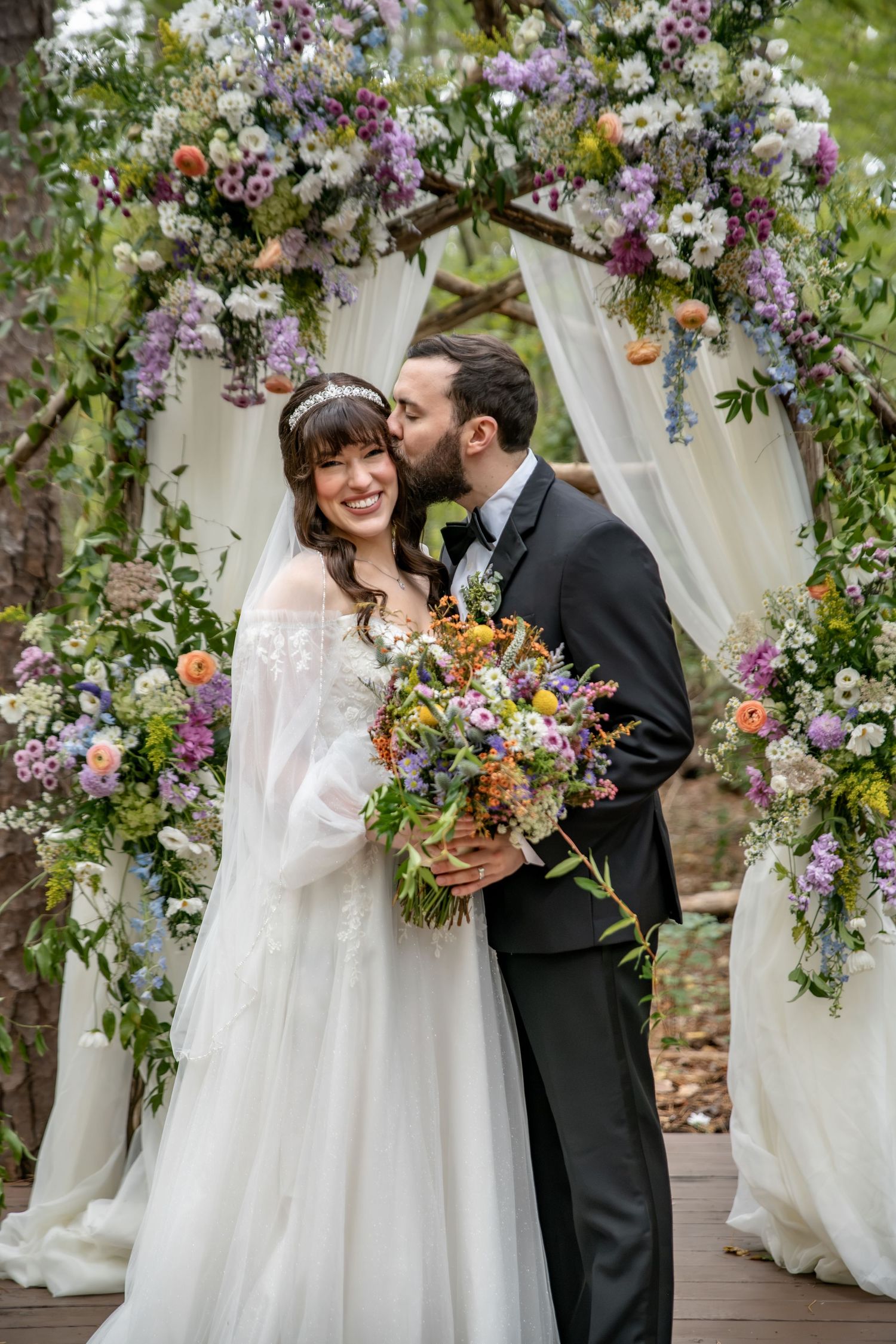Smiling bride in a flowing white gown holds a colorful bouquet as the groom kisses her cheek beneath a floral wedding arch.