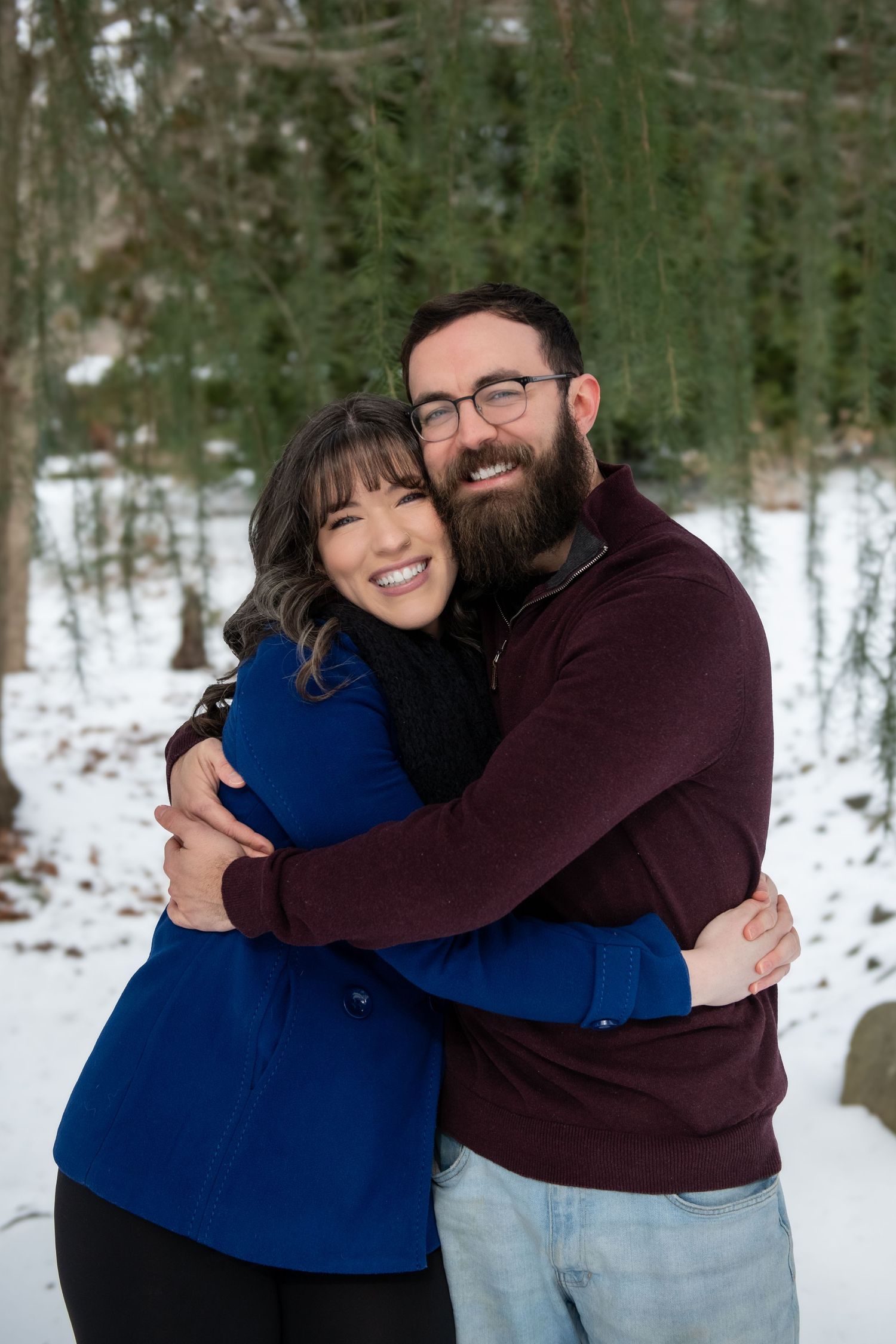 A couple shares warm embraces and laughter in a snowy winter setting with trees in the background.