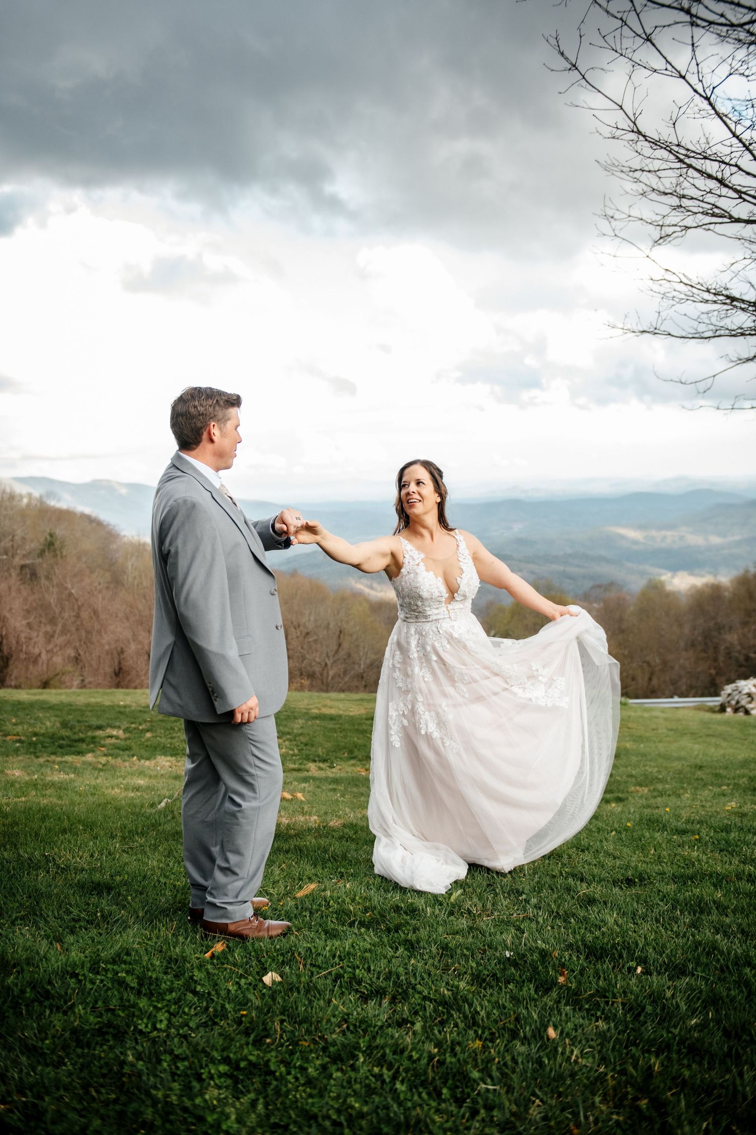 Bride and groom share a romantic dance on a green hillside with stunning mountain views under a cloudy sky.