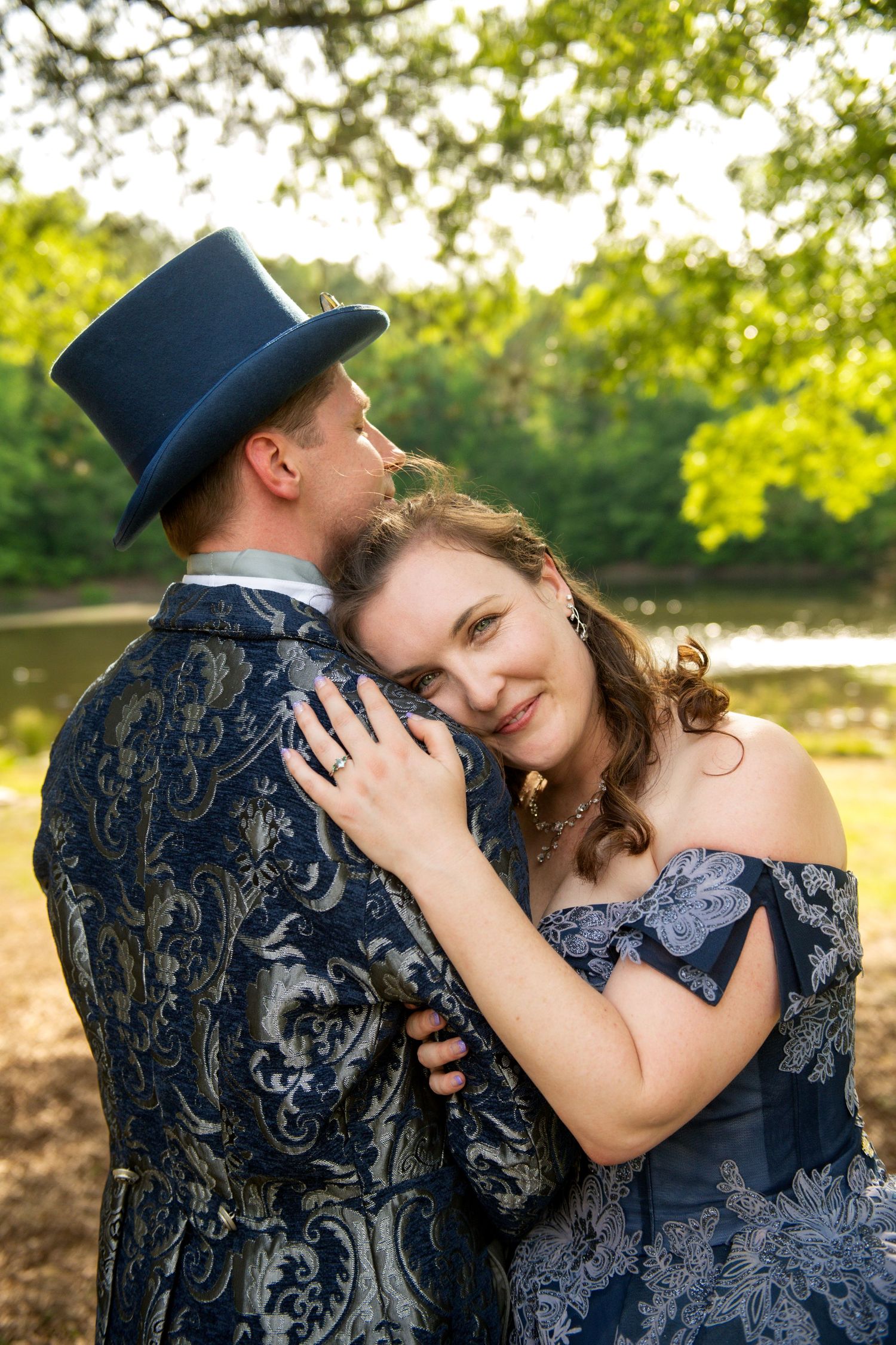 A couple in elegant blue Victorian attire embraces outdoors, the woman smiling warmly as the man wears a stylish top hat.