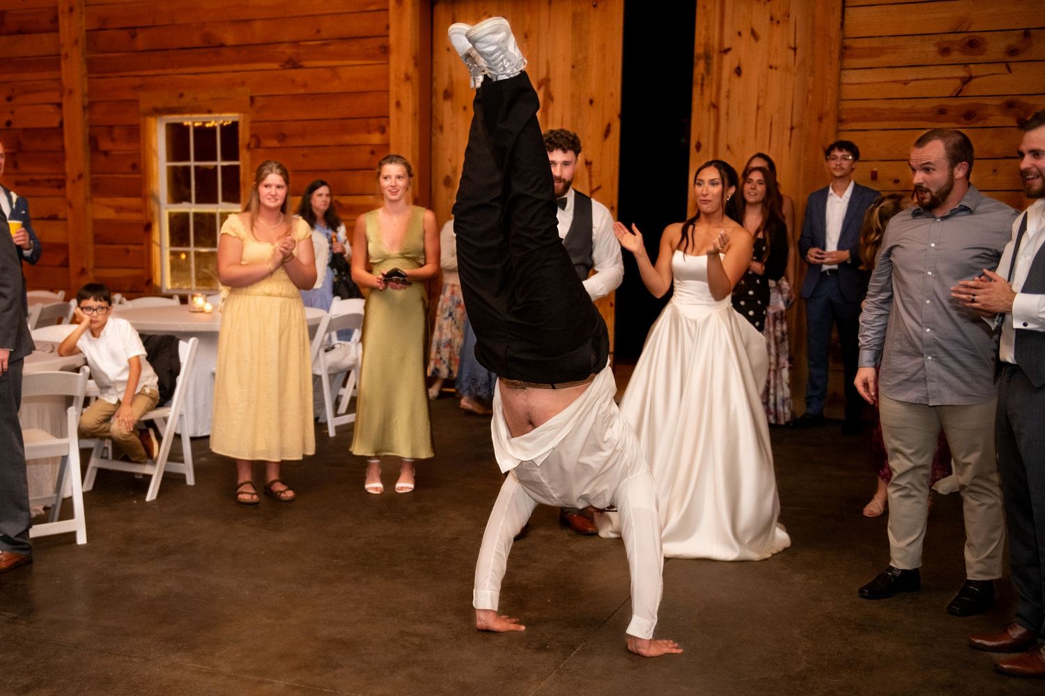 Groom doing a handstand at wedding reception while bride and guests watch and clap in rustic barn venue.