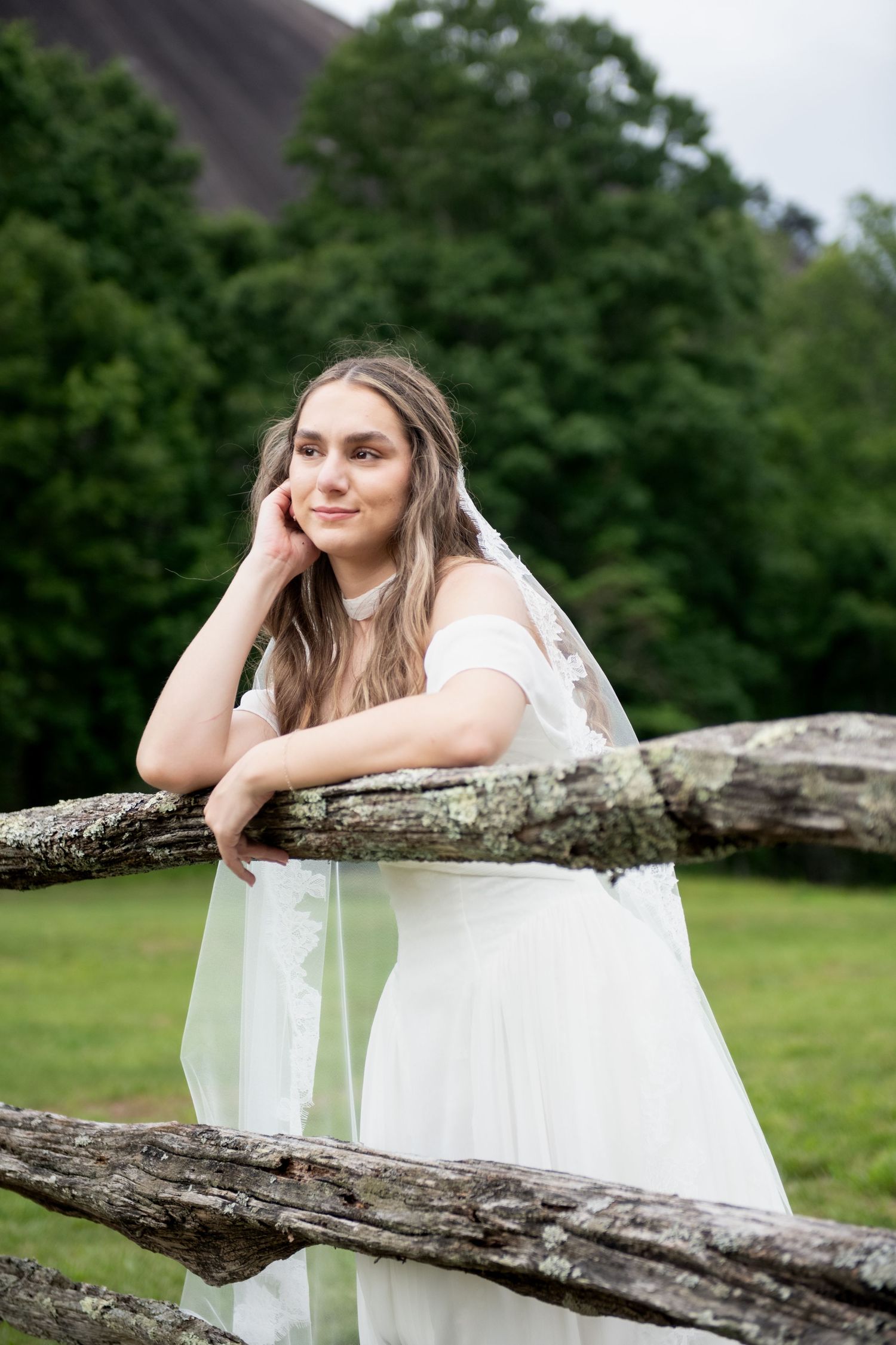 Bride in white gown and veil leans on rustic wooden fence, smiling dreamily with green trees in background.