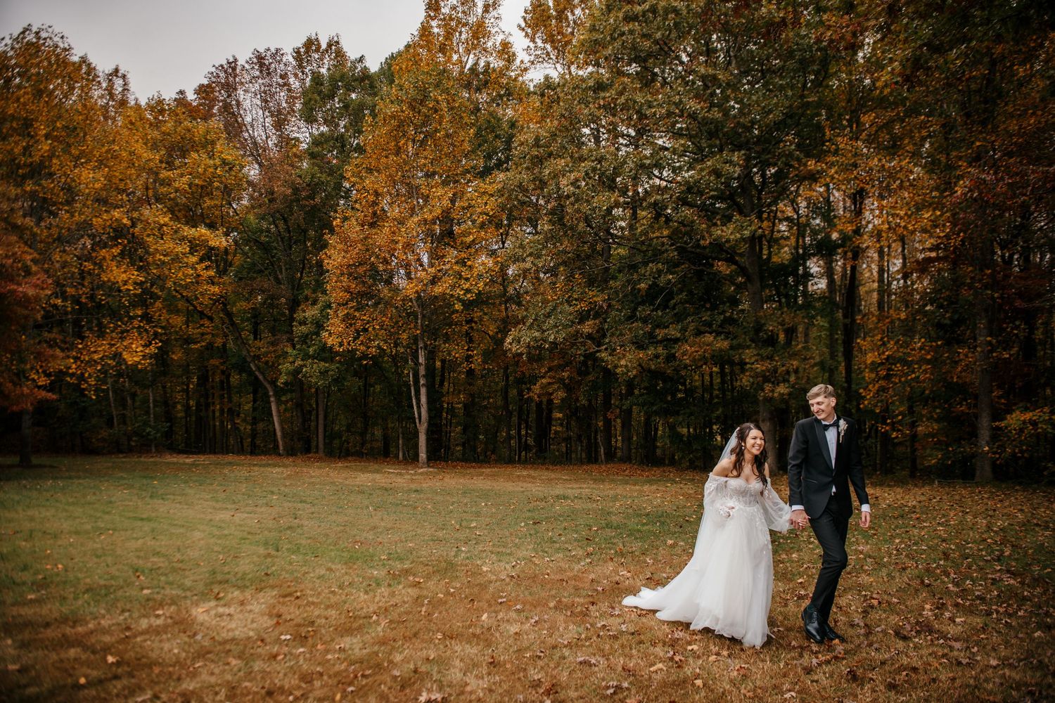A couple walks hand in hand through a meadow surrounded by trees with vibrant autumn foliage.