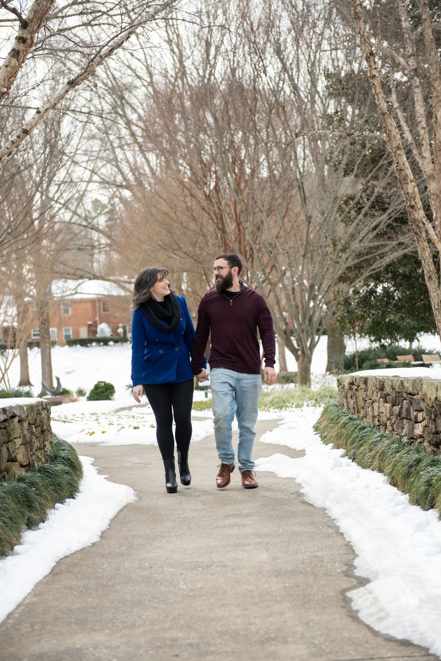 A couple takes a romantic winter stroll along a snowy path lined with bare trees.