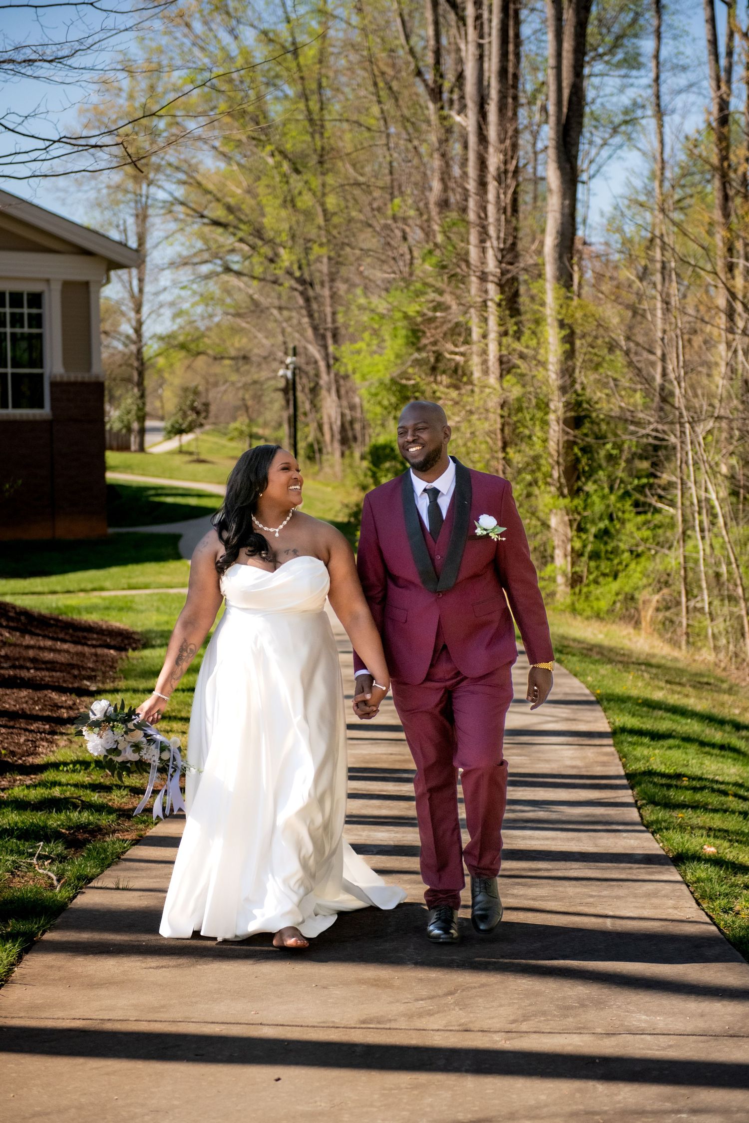 A bride and groom walk hand-in-hand on a sunlit path, she in white gown, he in burgundy suit, surrounded by spring trees.