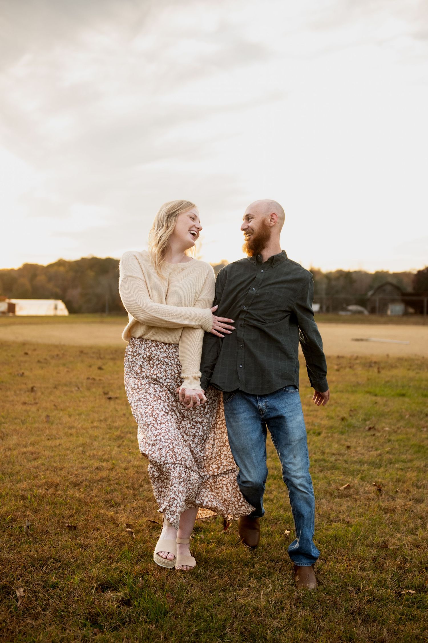 A couple laughs together while walking in an open field during golden hour sunset.